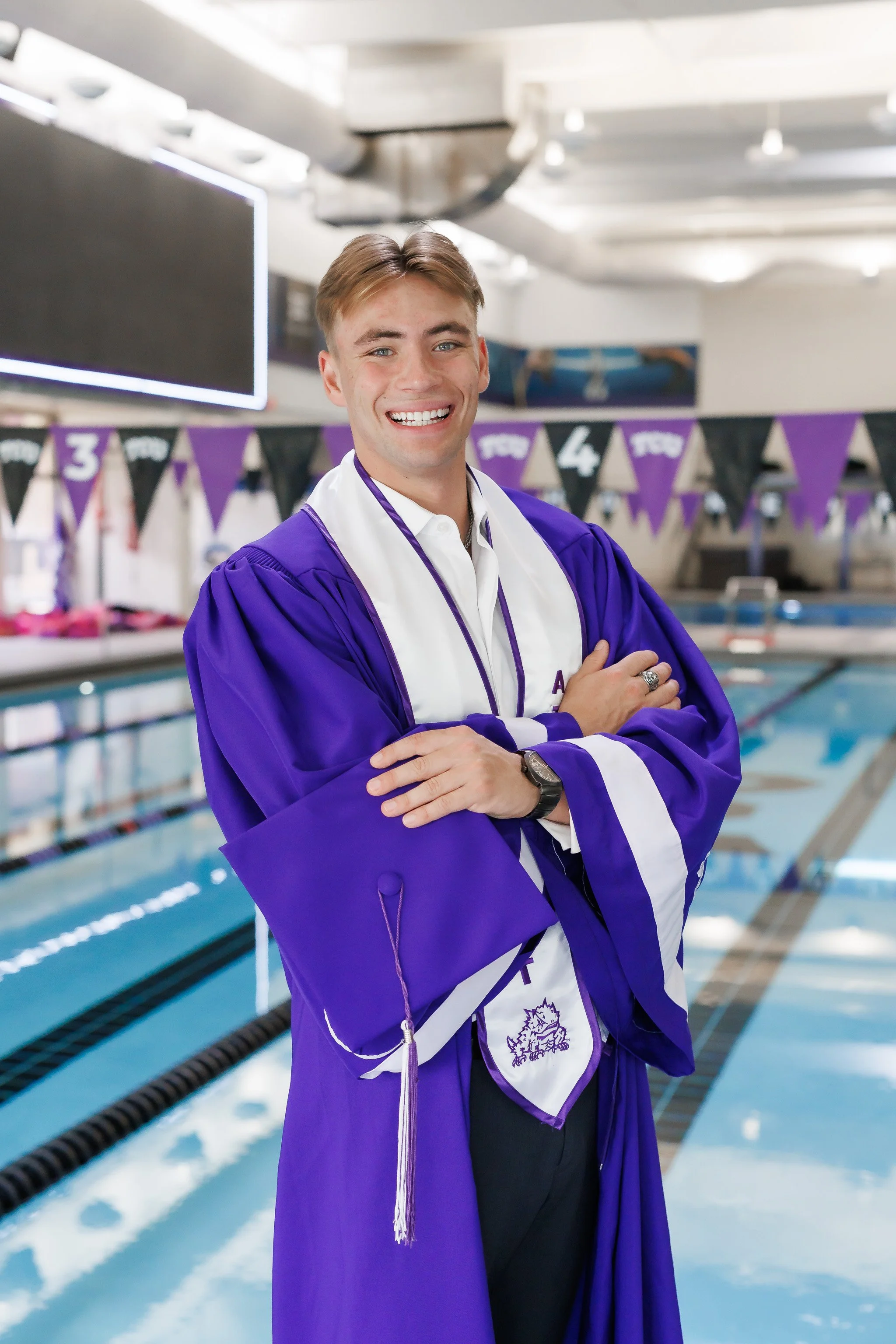 TCU Senior guy wearing a purple graduation gown, student athlete stole, holding a grad cap while standing at the side of the pool at the University Recreation Center posing for senior pictures