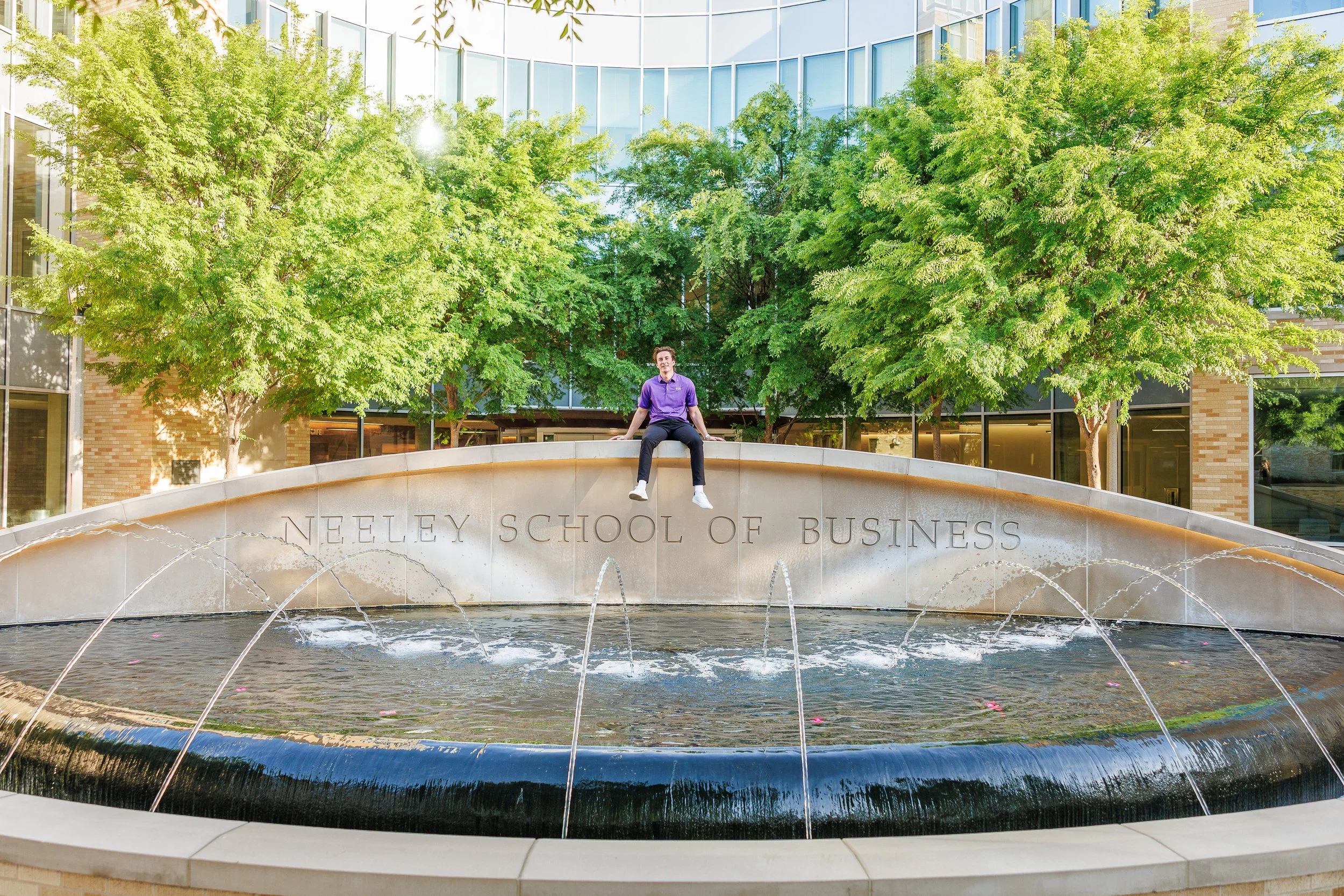 TCU senior guy sitting atop the water fountain at Neeley School of Business for Fort Worth graduation portraits