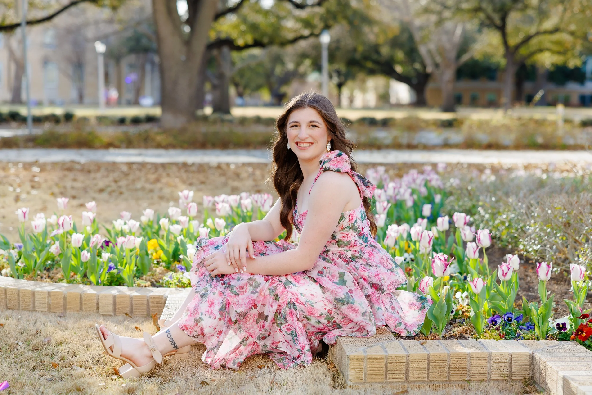 Senior girl at TCU wearing a pink floral dress sitting beside spring tulips in front of the TCU library