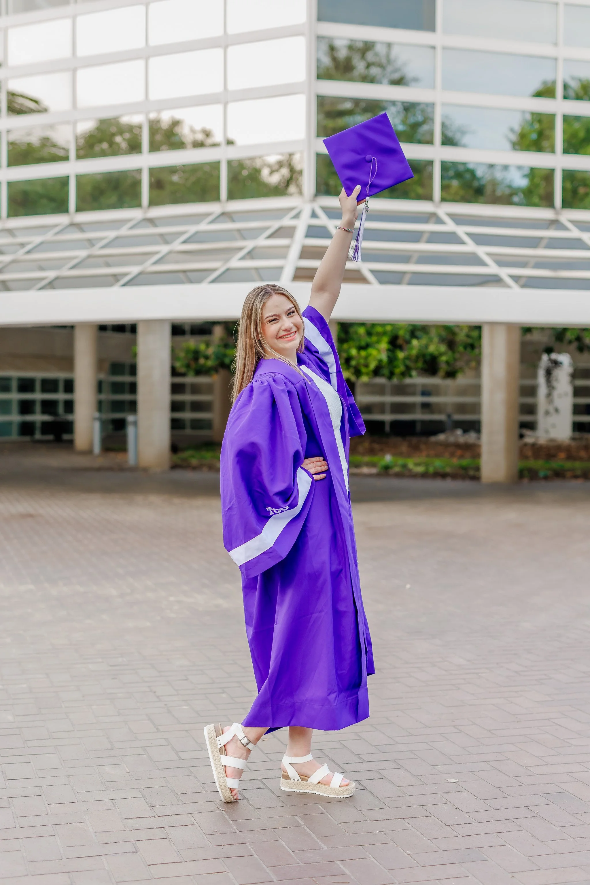 Senior girl wearing a purple graduation gown and waving a graduation cap in front of the Bob Schieffer College of Communications at TCU posing for senior pictures