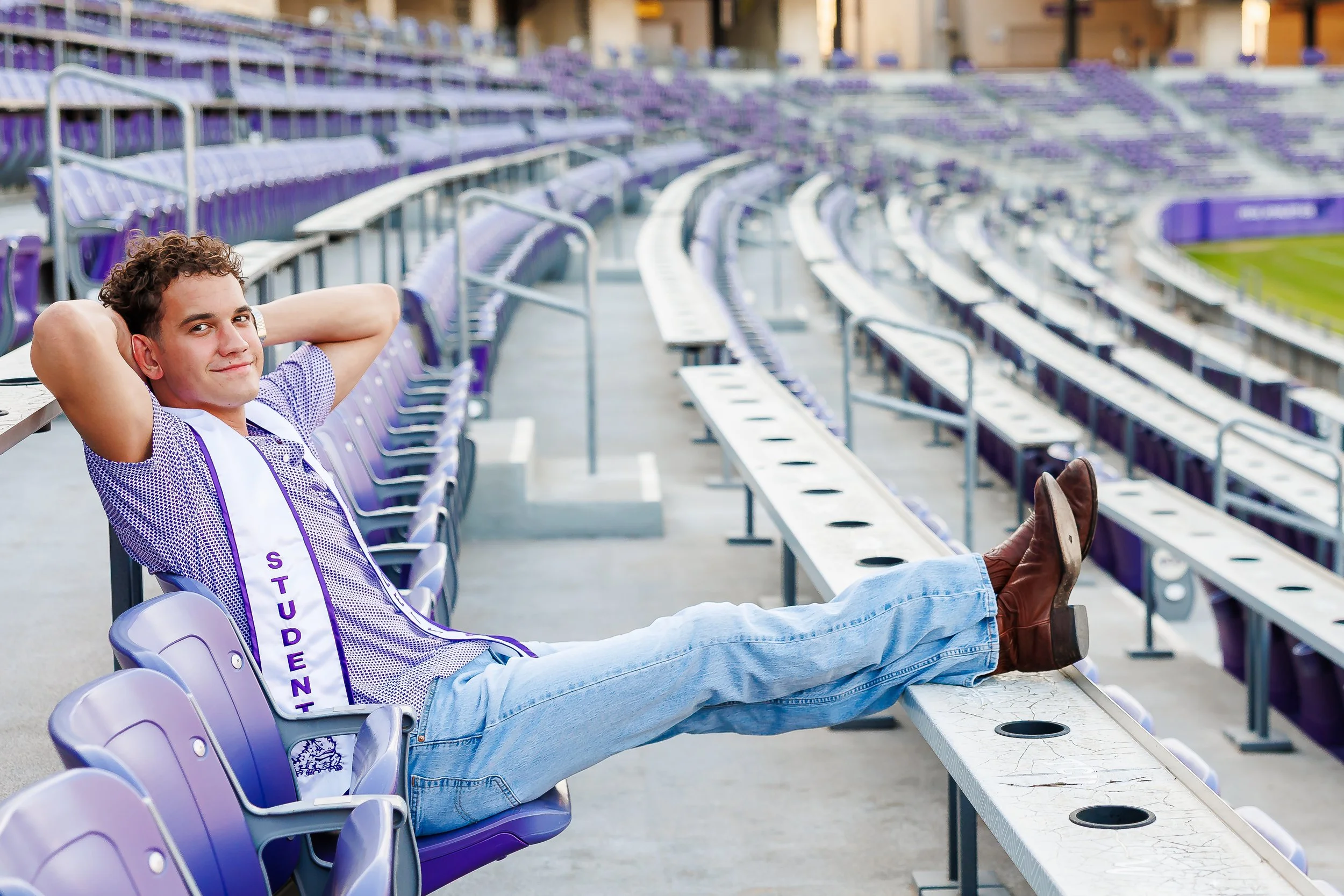 A relaxed TCU graduate lounges in the stadium seats with boots up and hands behind his head, wearing a “Student Athlete” stole. A casual and confident Dallas graduation photo that captures school pride and personality
