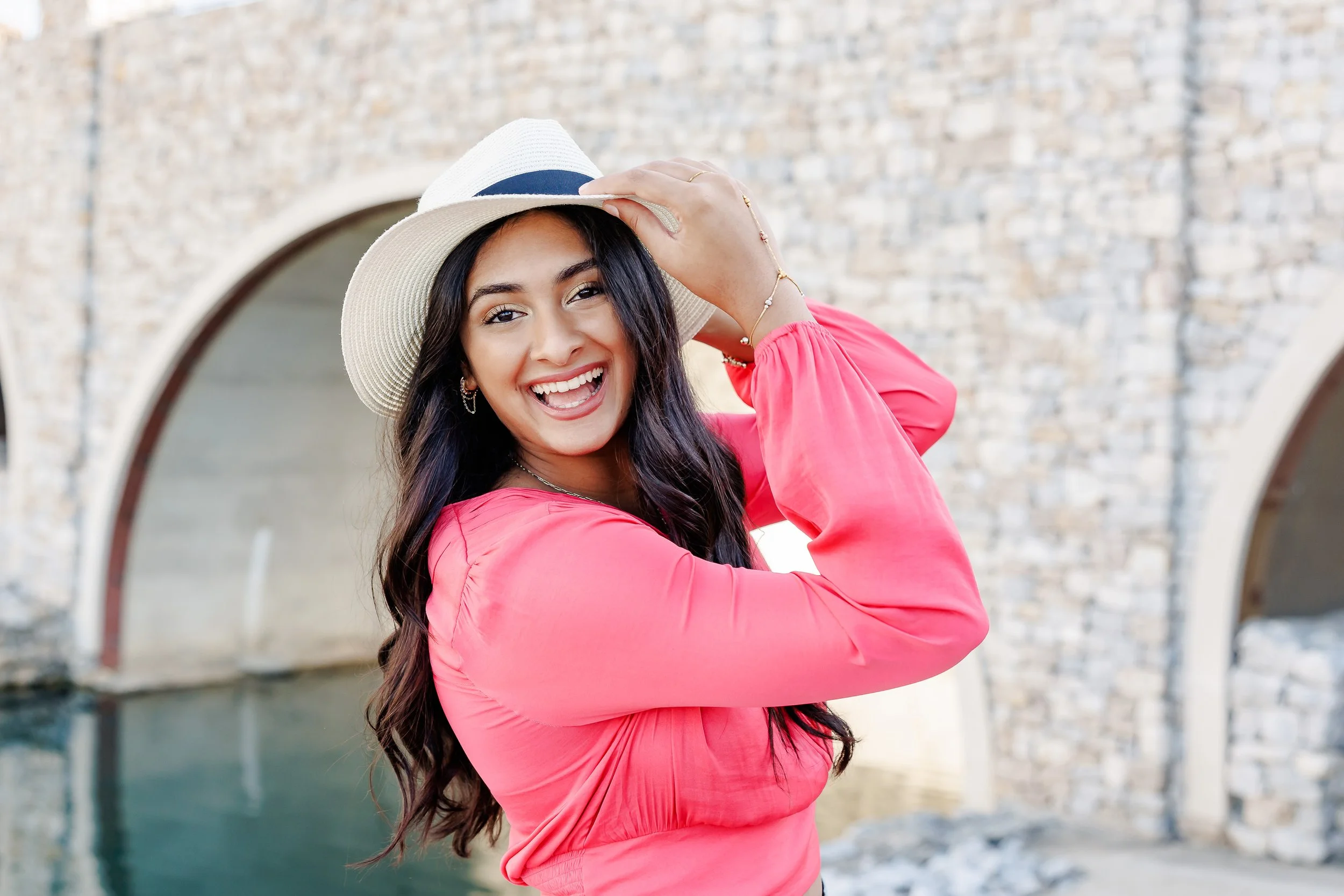 young lady with long black hair wearing a hot pink blouse and white straw hat laughing at the camera in front of a water canal and bridge posing for professional portraits Westlake Entrada