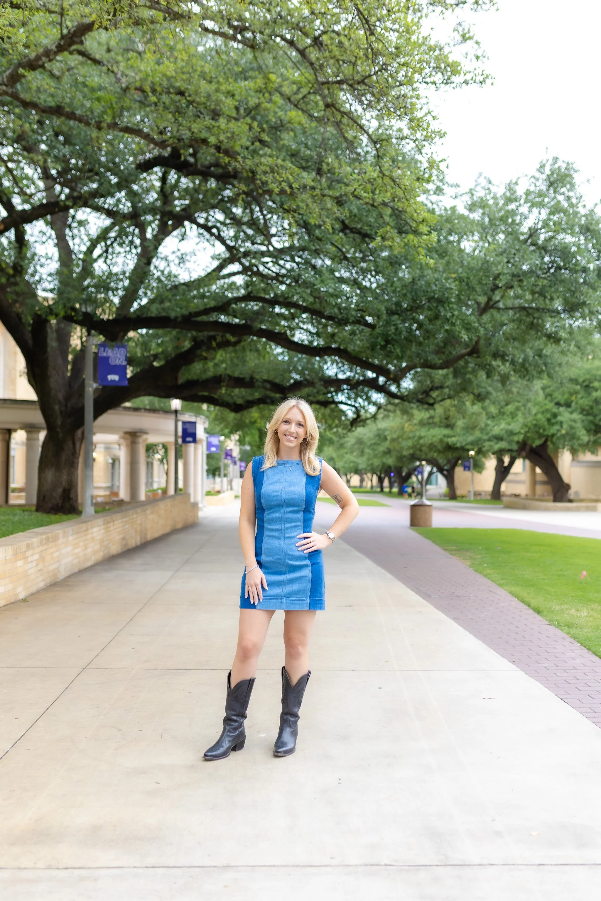 Senior girl wearing a blue denim dress and brown cowboy boots standing outside of the TCU library posing for senior pictures