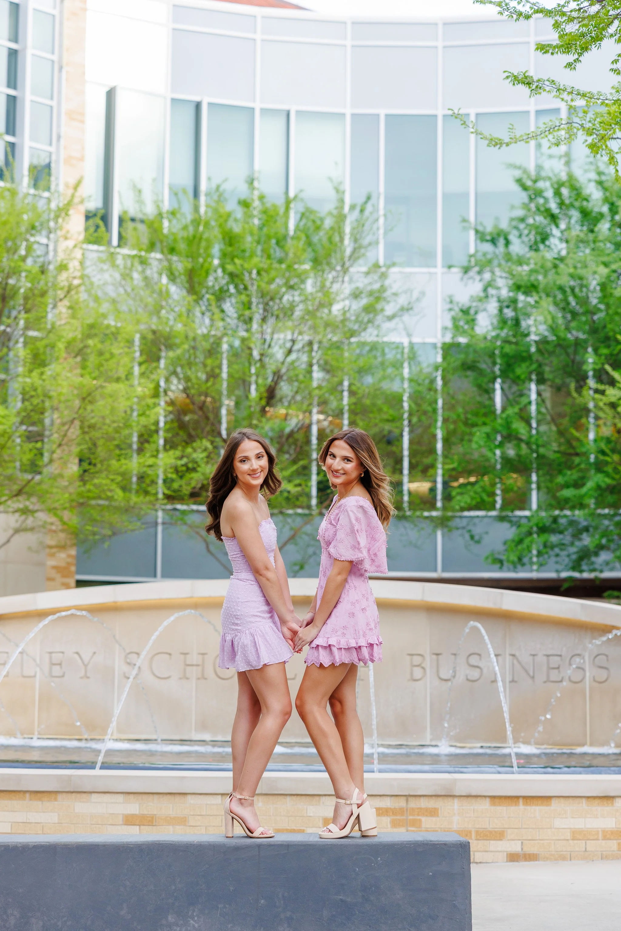 TCU twin senior girls wearing lavender dresses holding hands in front of the water fountain at Neeley School of Business posing for senior pictures
