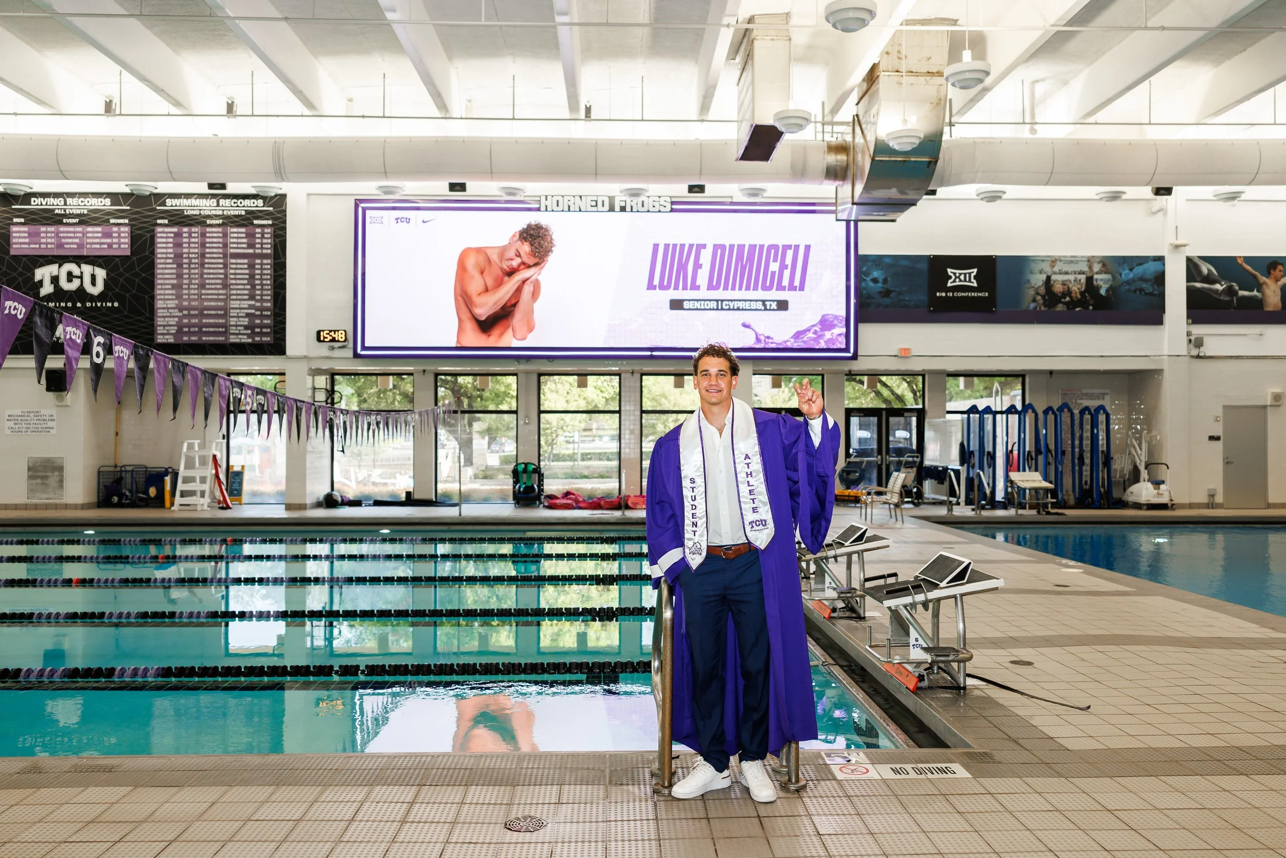 TCU Senior guy wearing a purple graduation gown and student athlete stolestanding at the side of the pool at the University Recreation Center posing for senior pictures