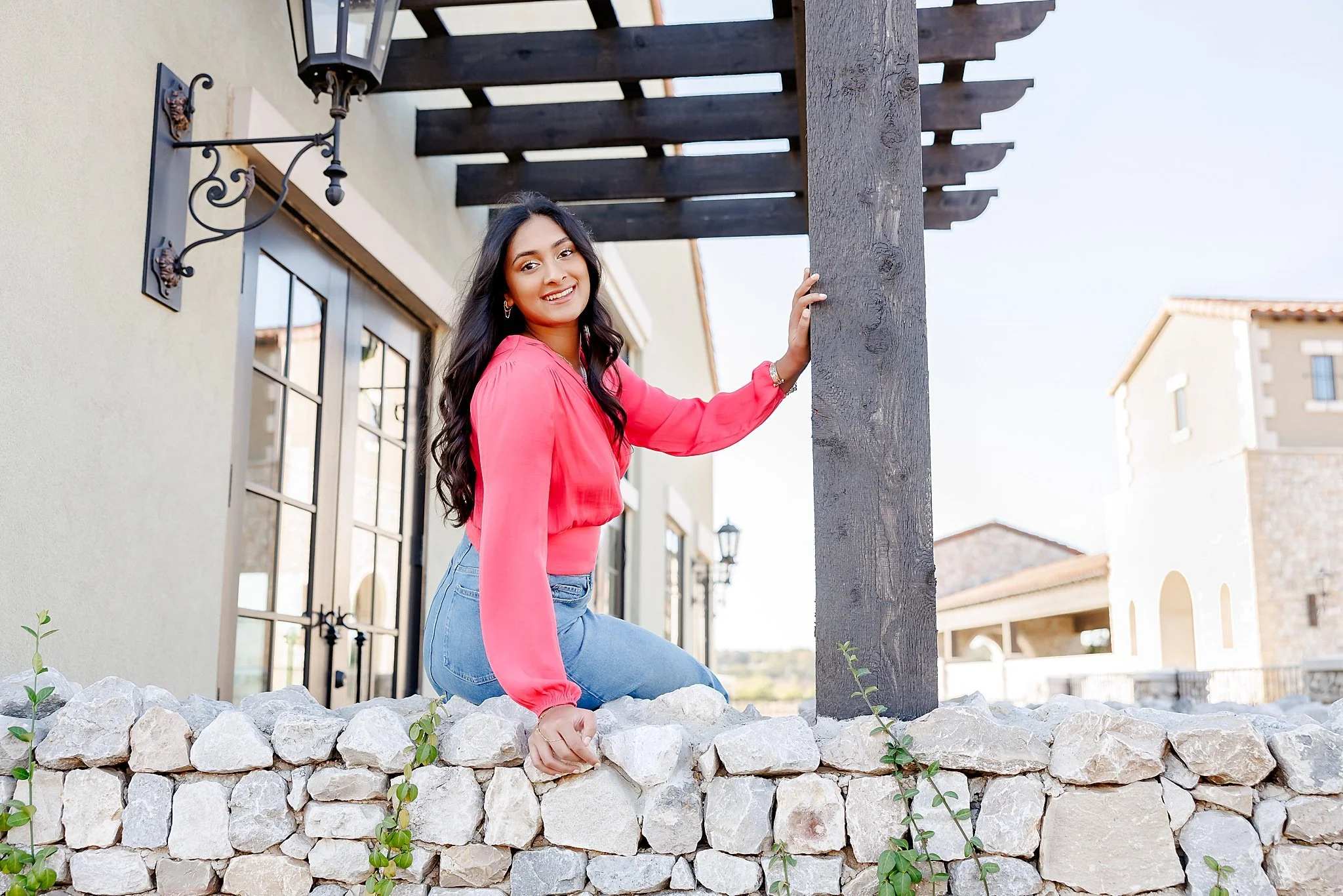 young lady with long black hair wearing hot pink blouse and long denim skirt sitting on a rock wall smiling at the camera posing for senior pictures at Westlake Entrada with DeAndra Jarboe Photography