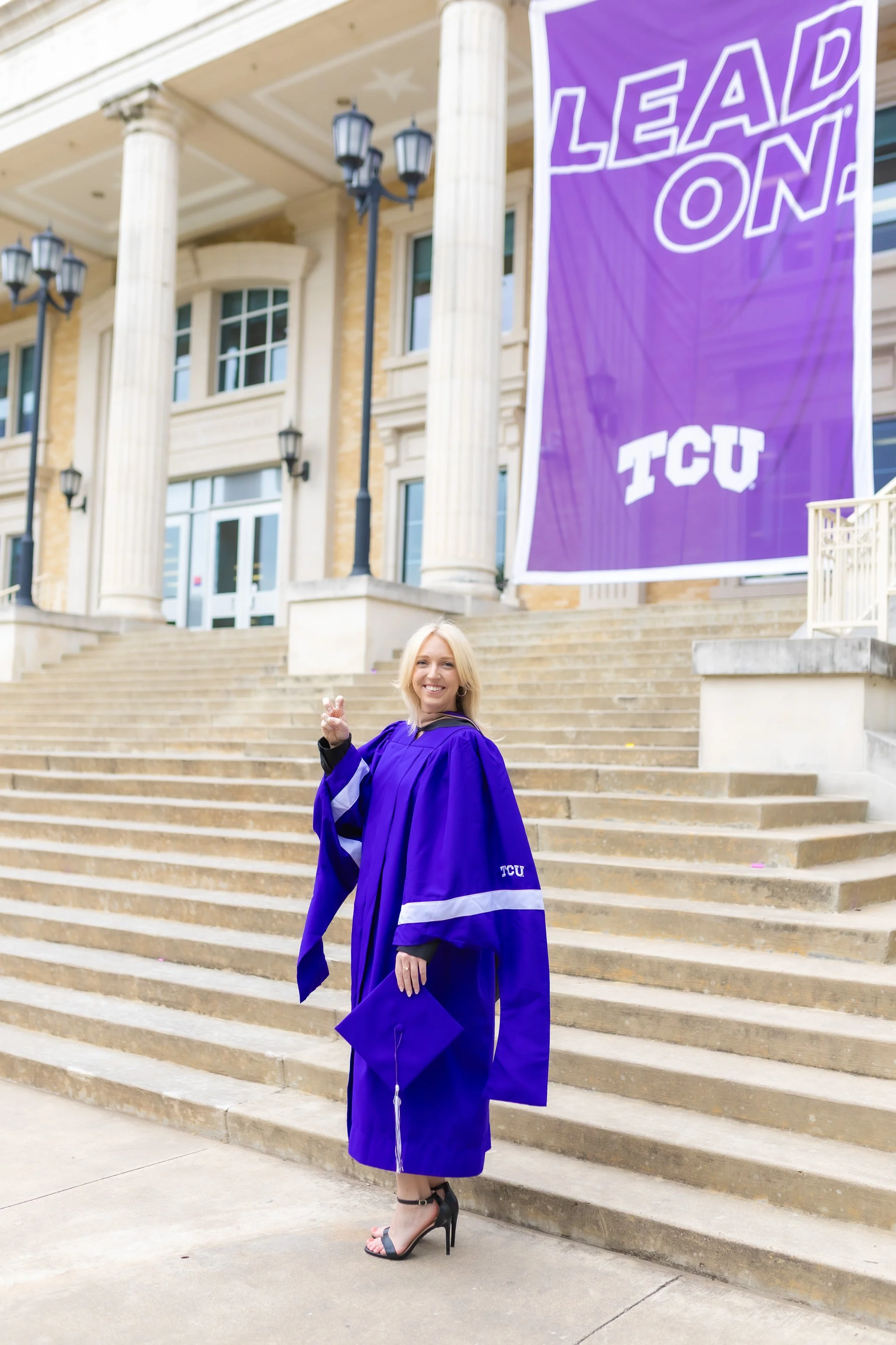 Senior girl wearing a purple graduation cap and gown nearthe steps of the TCU library posing for senior pictures
