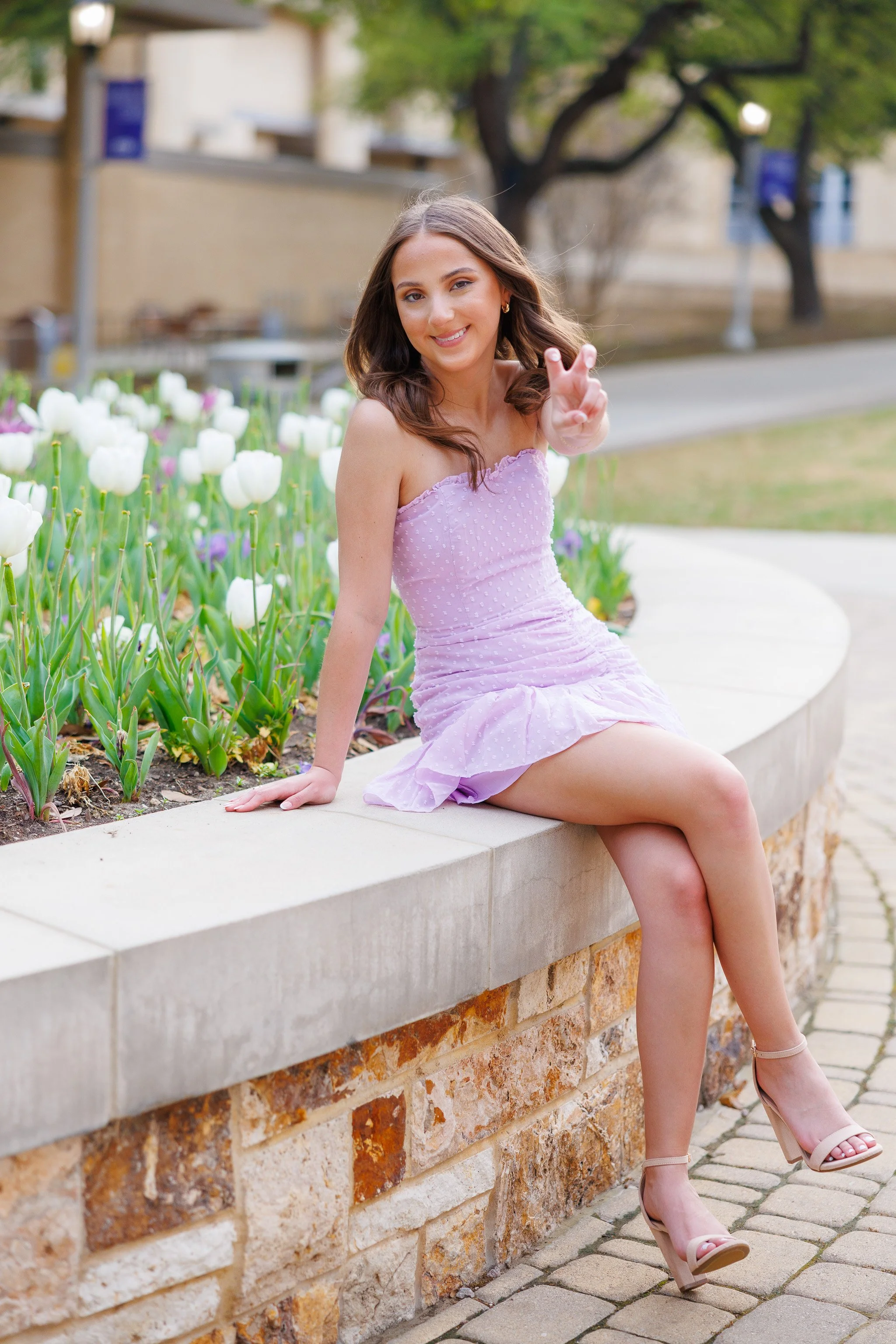 Senior girl wearing a short lavender dress sitting next to the spring tulips outside near the TCU library posing for senior pictures