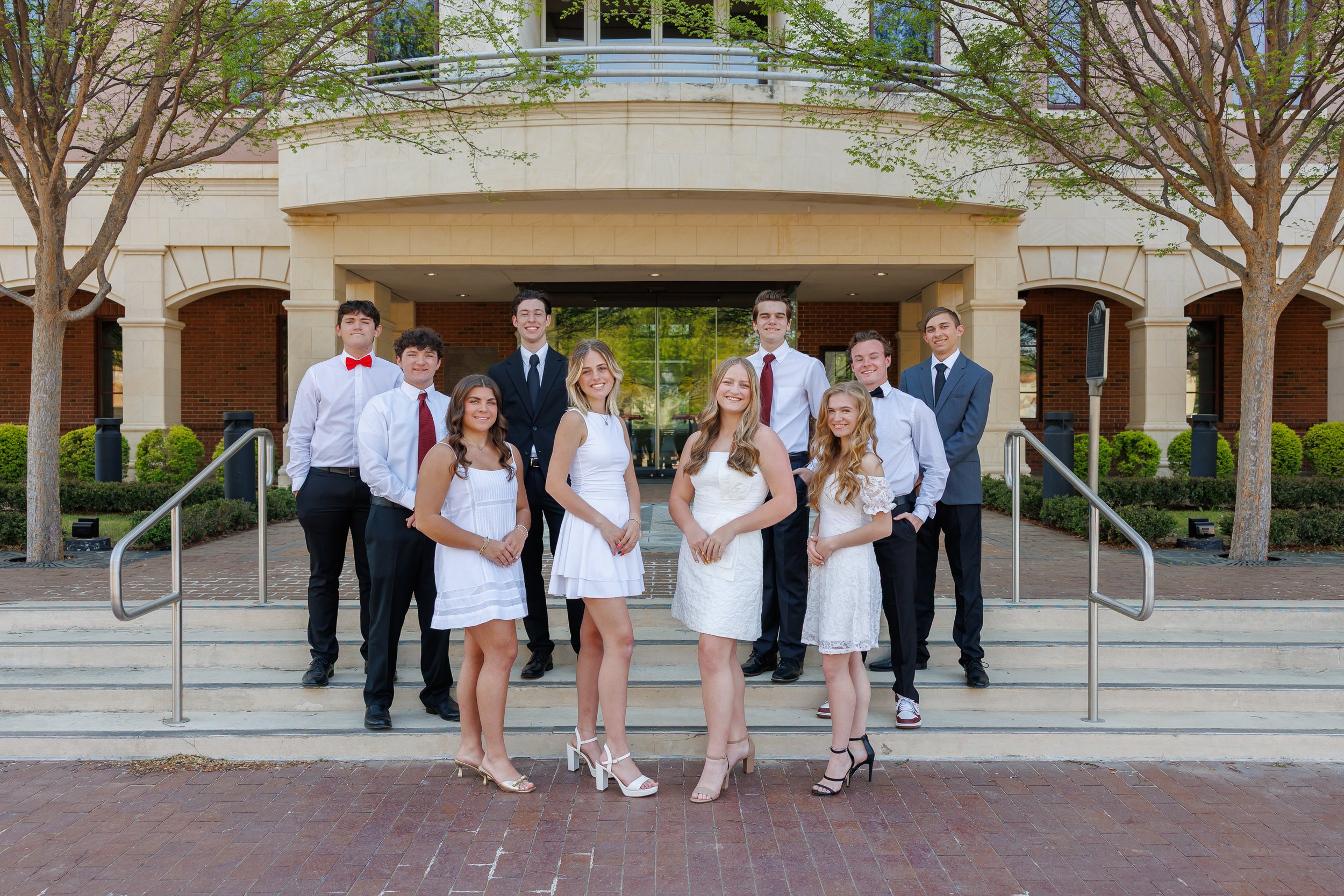 group of ten highschool senior students posing for class pictures standing on the steps of Colleyville Library. Girls are wearing white dresses and heels. Guys wearing dress pants with white button down shirts.  Everyone is smiling at the camera.