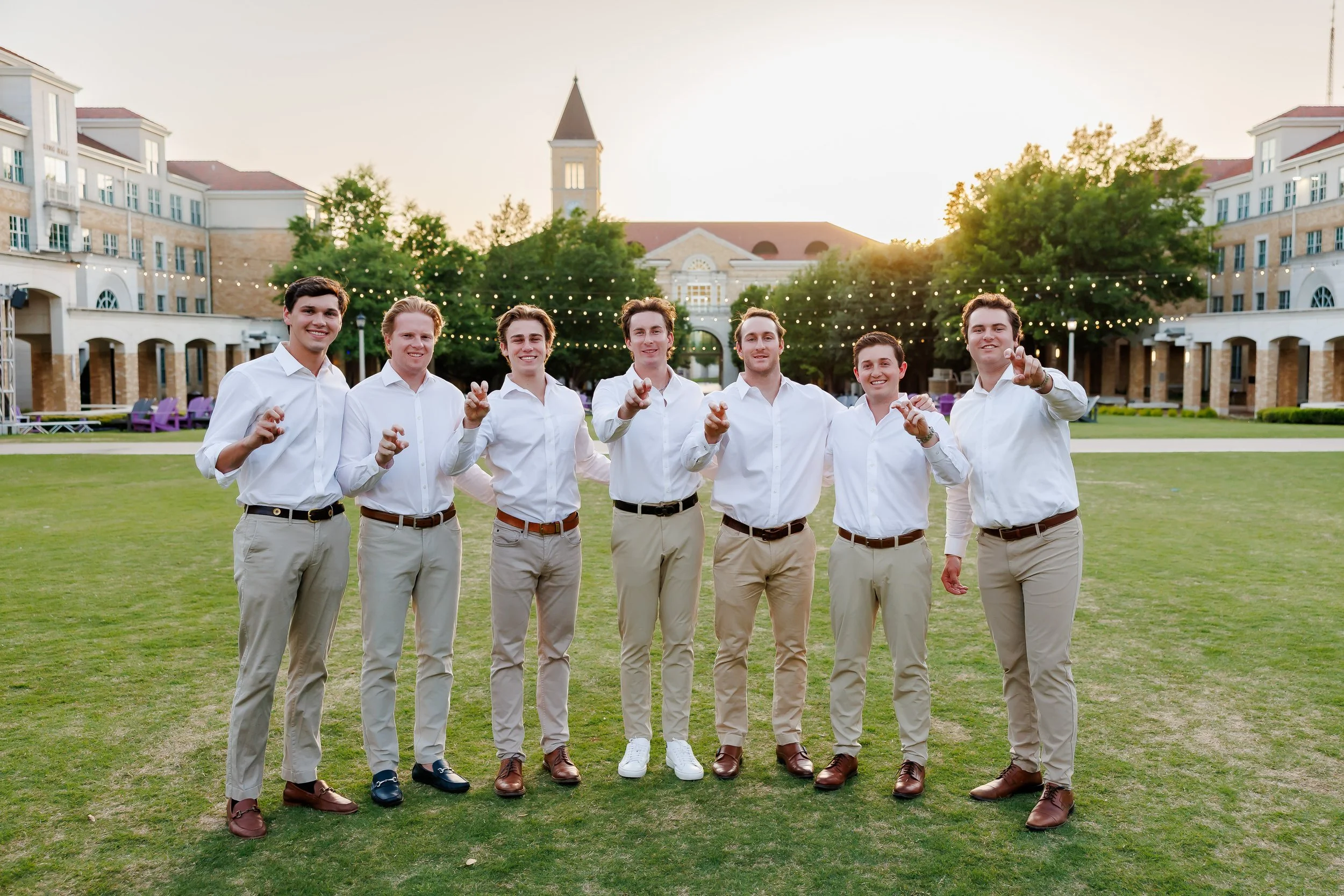 Group of seven TCU senior guys wearing white shirts and khaki pants standing and hugging on the lawn of The Commons