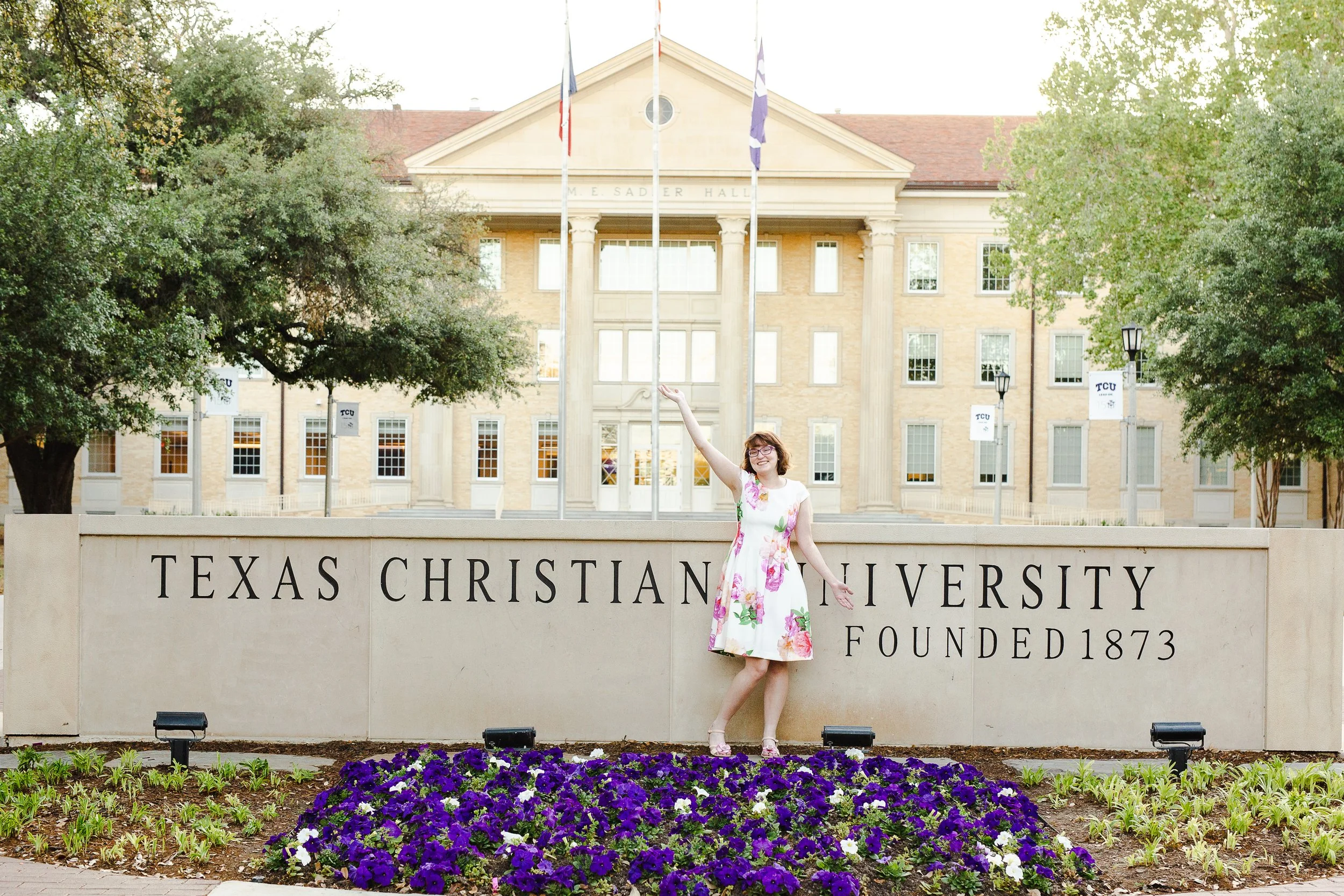 Graduate in a floral dress poses with one hand raised and the other resting on the Texas Christian University sign, celebrating in front of Sadler Hall.