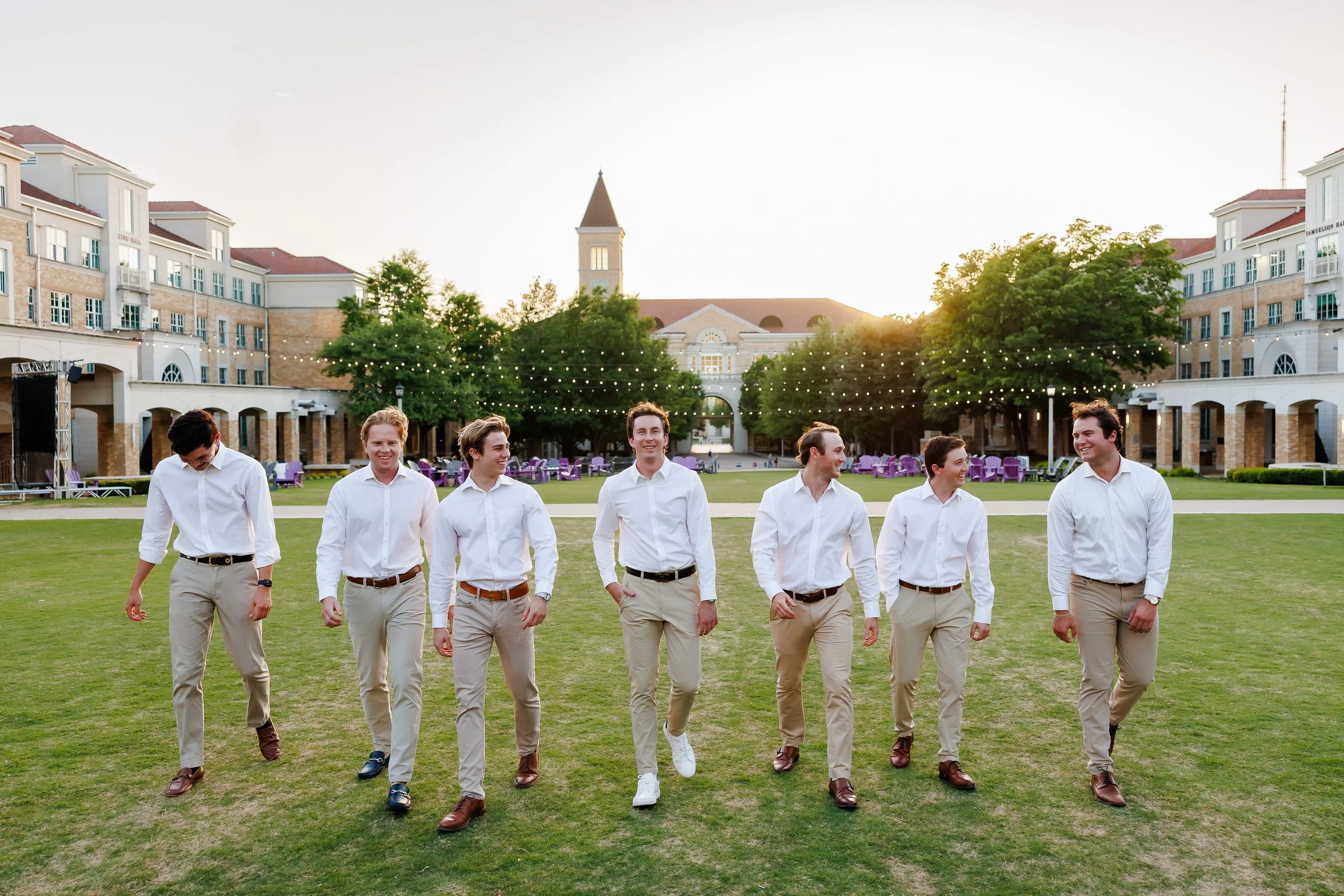Group of seven TCU senior guys wearing white shirts and khaki pants walking and laughing on the lawn of The Commons