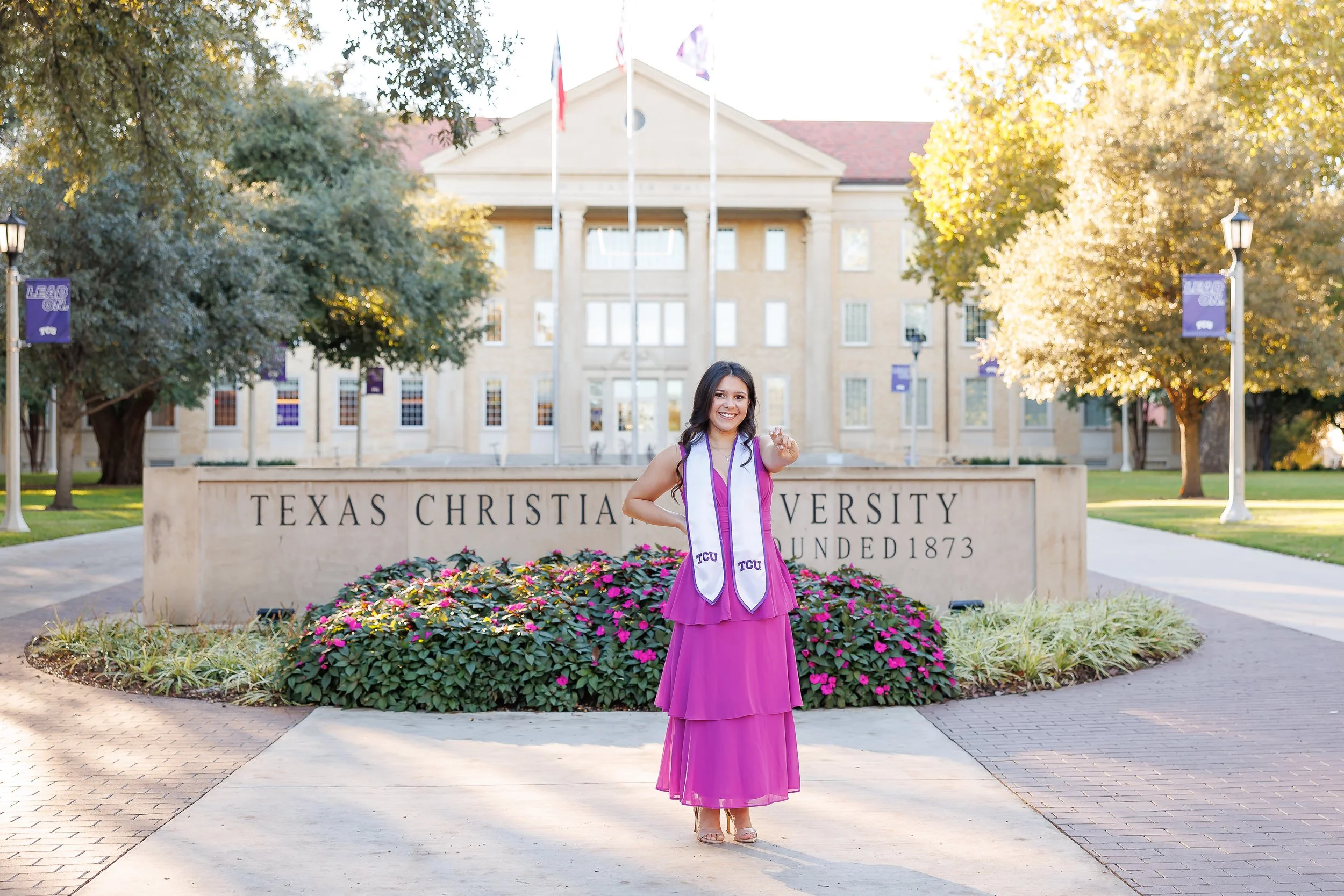 Graduating senior in a magenta dress and TCU stole poses in front of the Texas Christian University sign, making the Horned Frog hand gesture, photographed by a Dallas senior photographer.