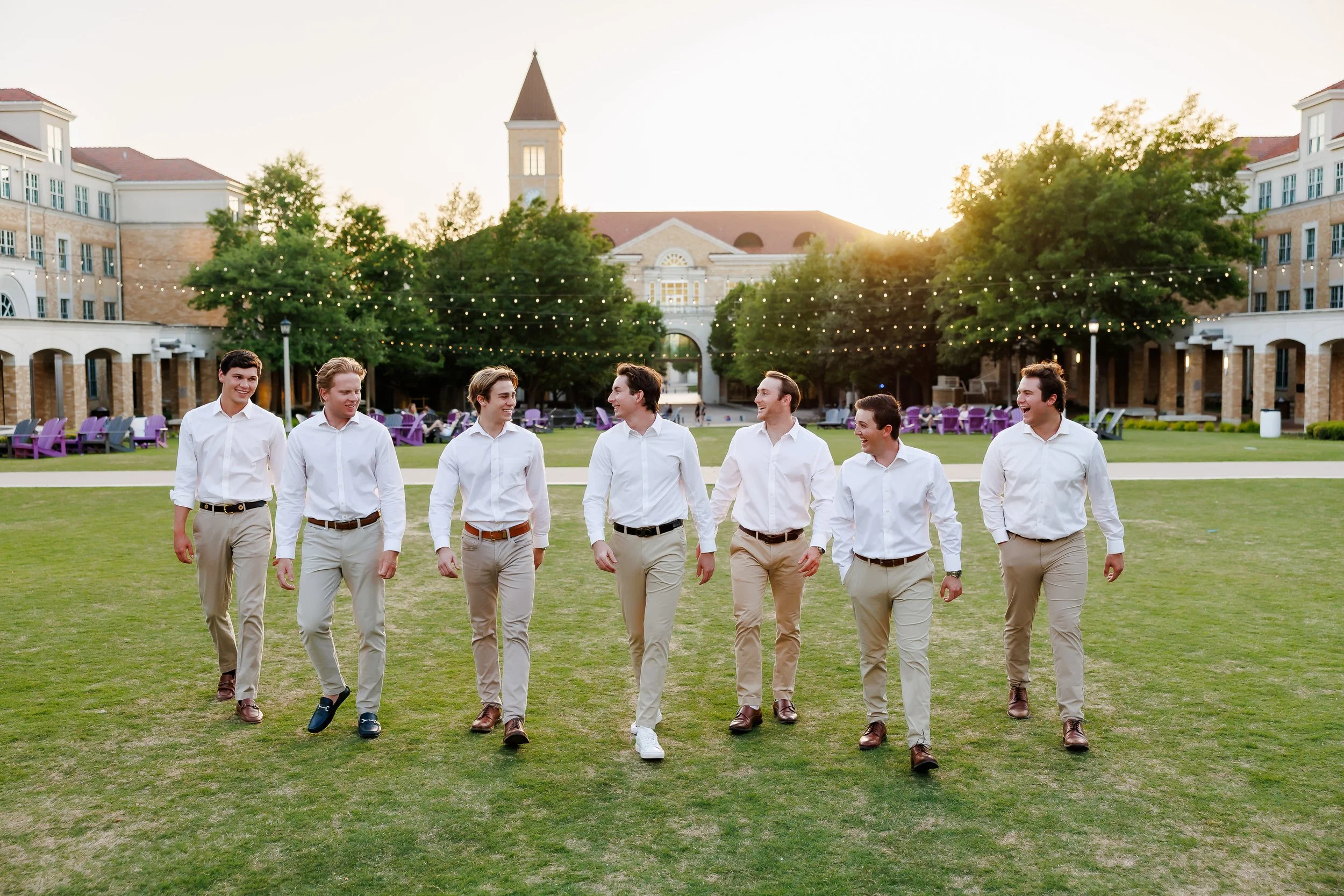 Group of seven TCU senior guys wearing white shirts and khaki pants walking and laughing on the lawn of The Commons