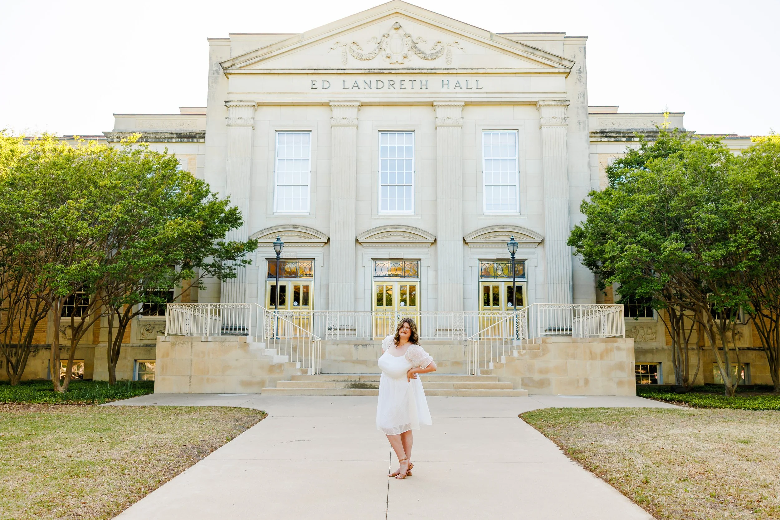 TCU senior girl in white dress waving a her hand outside the Ed Landreth Hall for graduation portraits