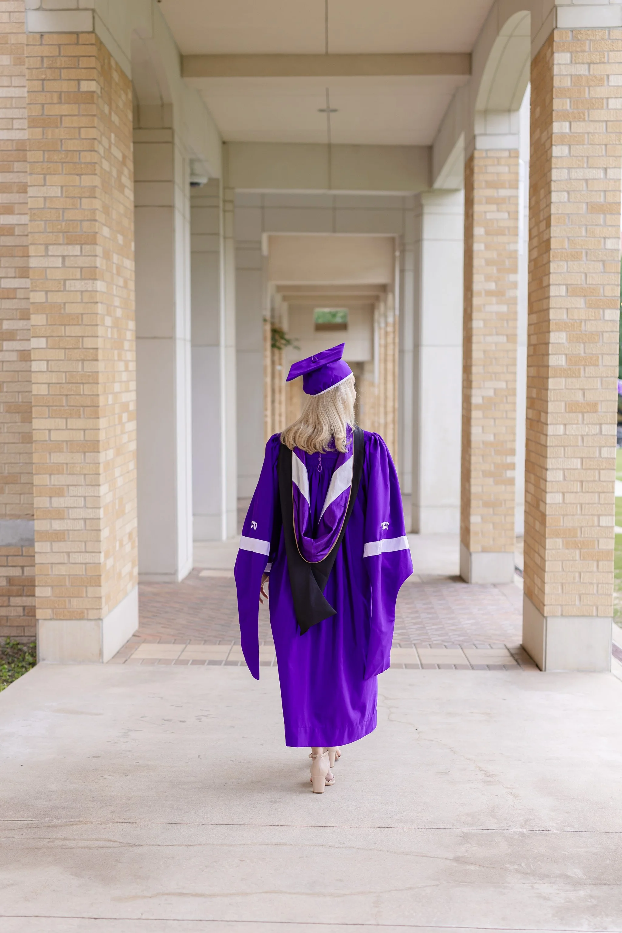 TCU graduate student in cap and gown with cords standing under the columns in The Commons for senior portraits