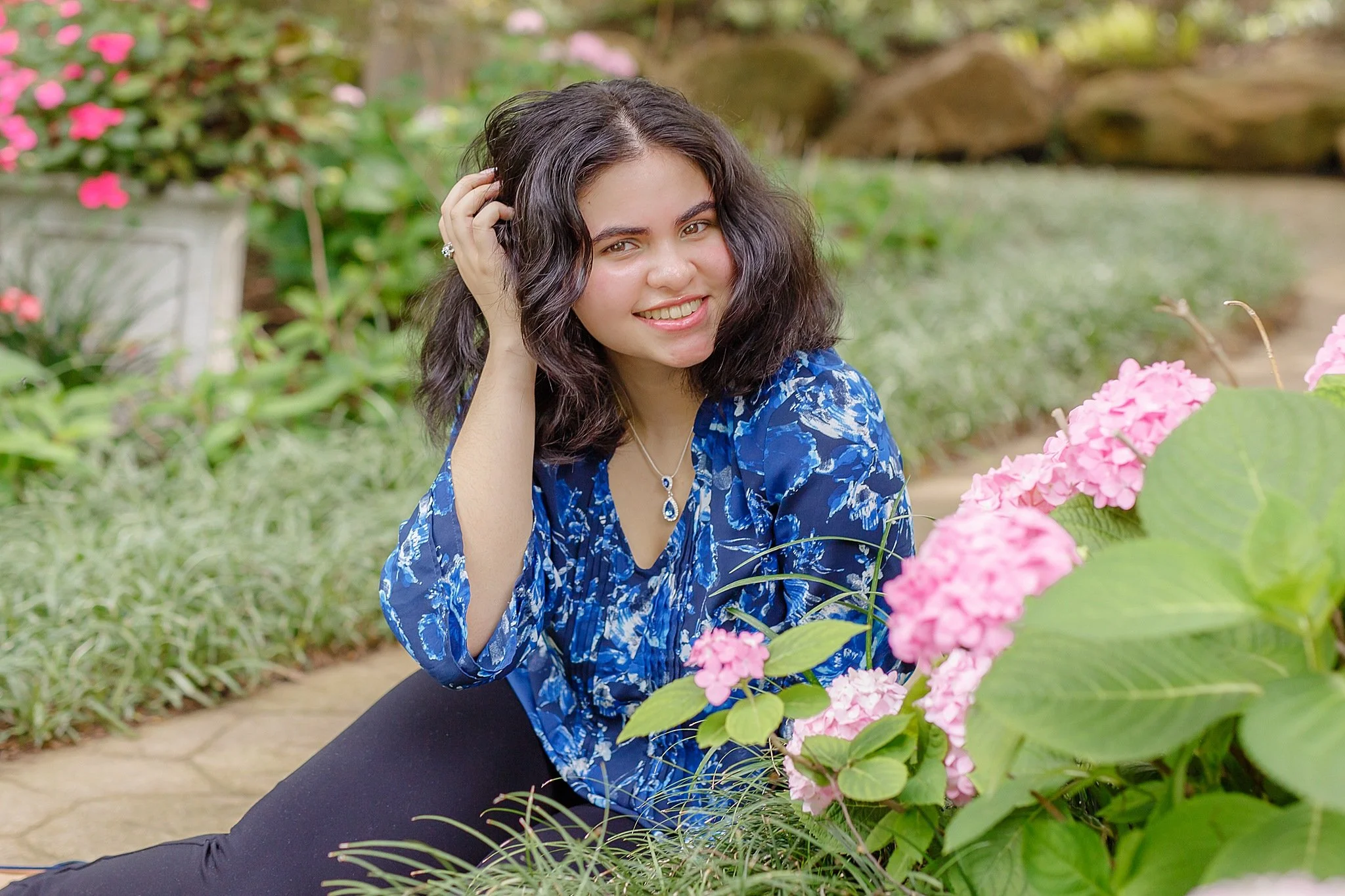 young lady with black wavy hair wearing blue floral blouse and navy pants smiling at the camera sitting on the ground posing for senior pictures at grapevine botanical gardens