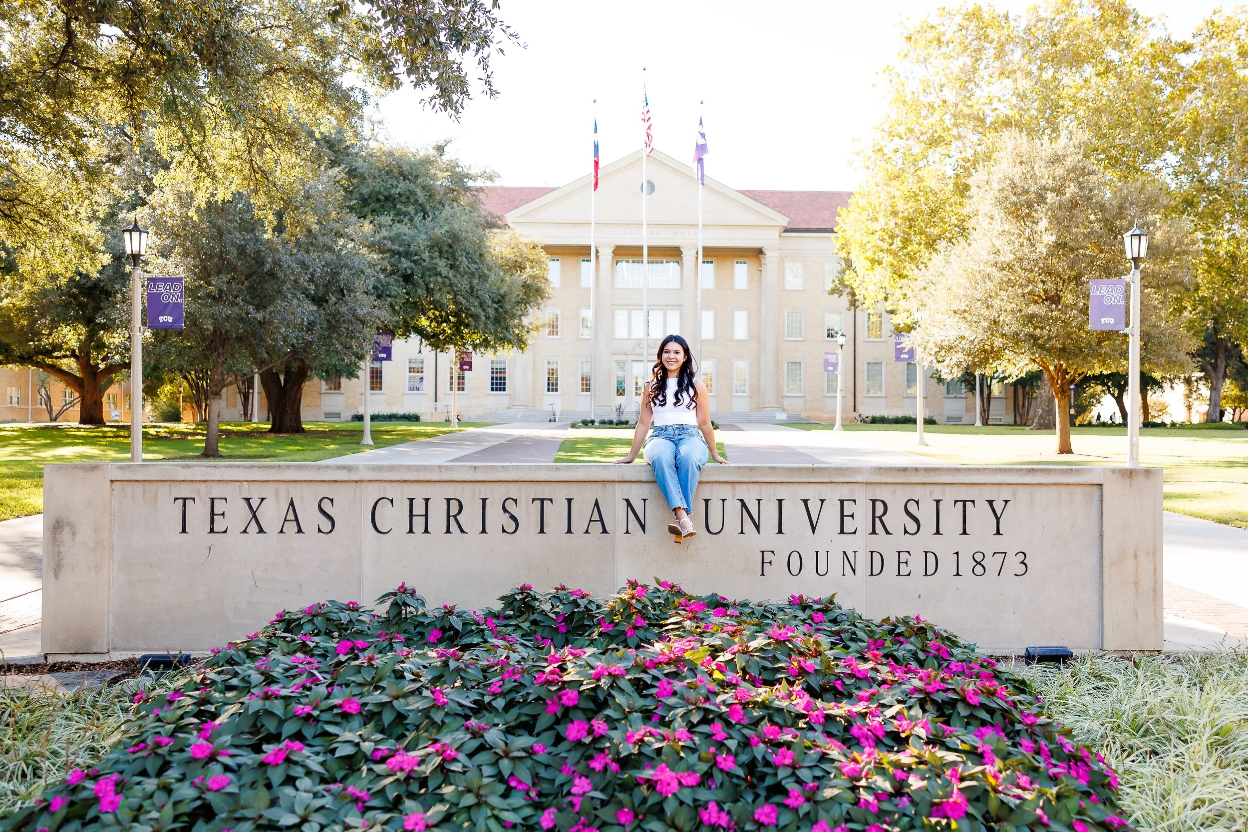 senior poses in front of the Texas Christian University sign on a sunny day, surrounded by blooming flowers and campus greenery, captured by a Dallas senior photographer
