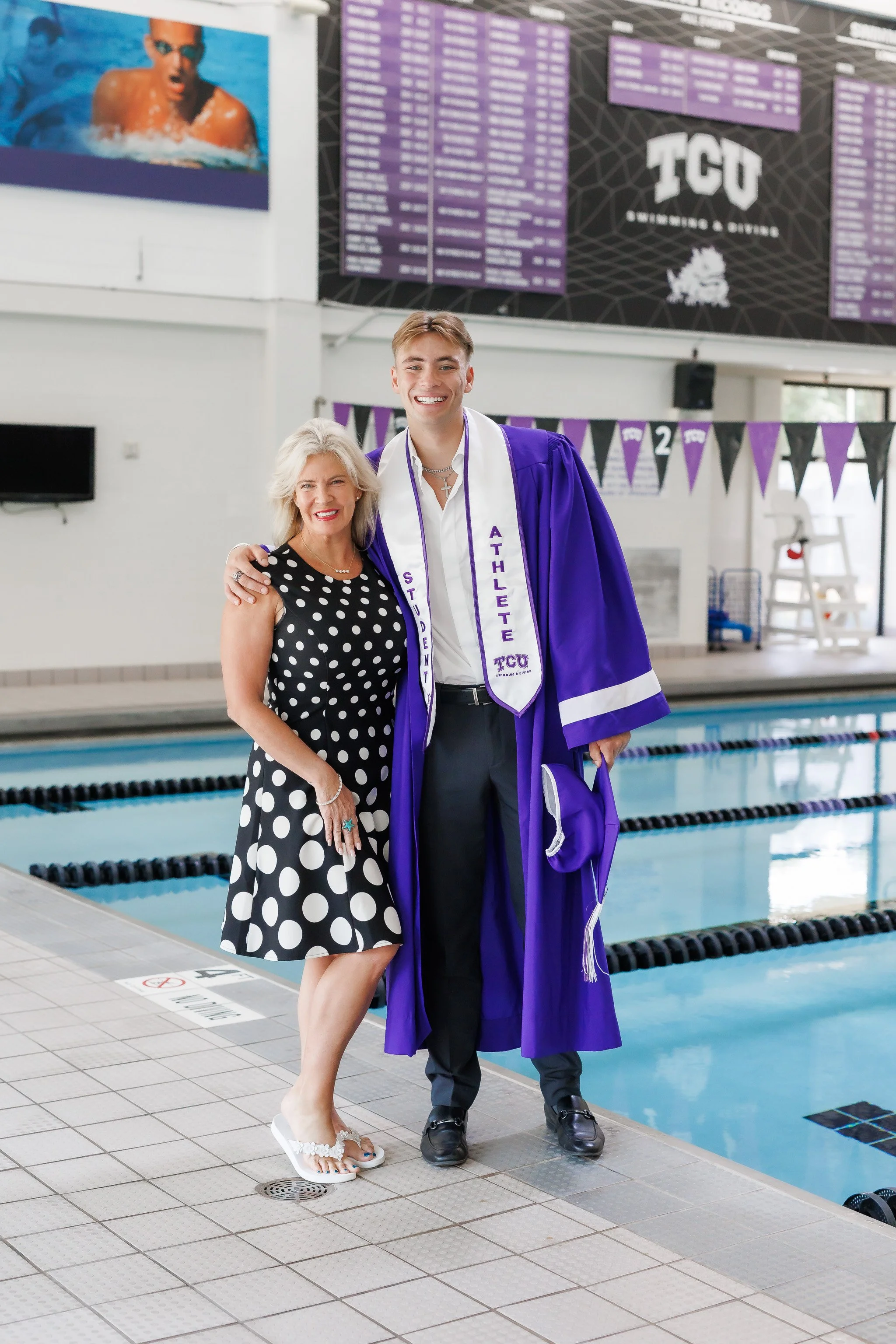 TCU Senior guy wearing a purple graduation gown, student athlete stole, holding a grad cap and hugging his mother while standing at the side of the pool at the University Recreation Center posing for senior pictures