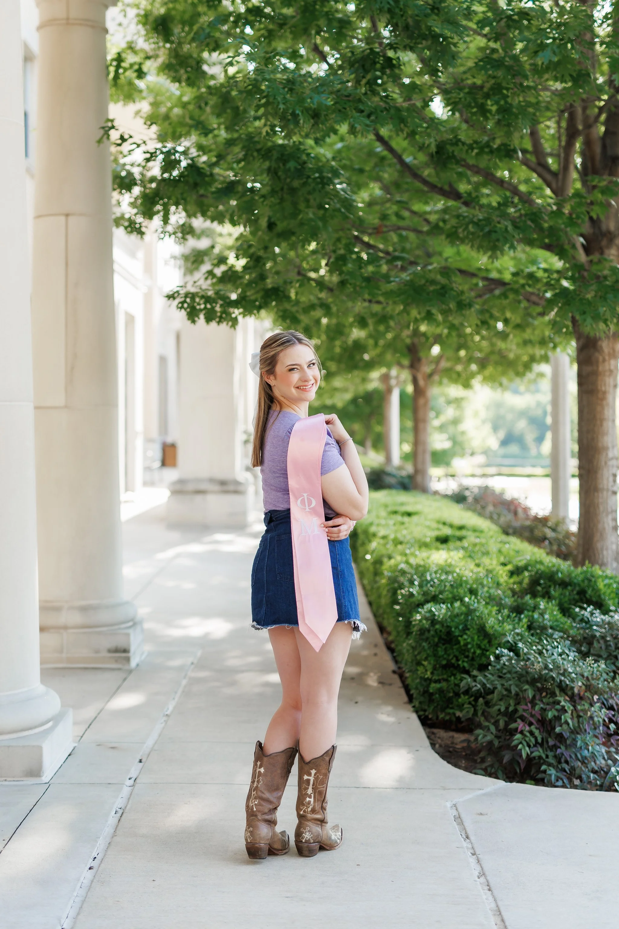 TCU senior girl wearing denim skirt lavender blouse brown boot holding a sorority stole in front of Phi Mu sorority house posing for senior pictures