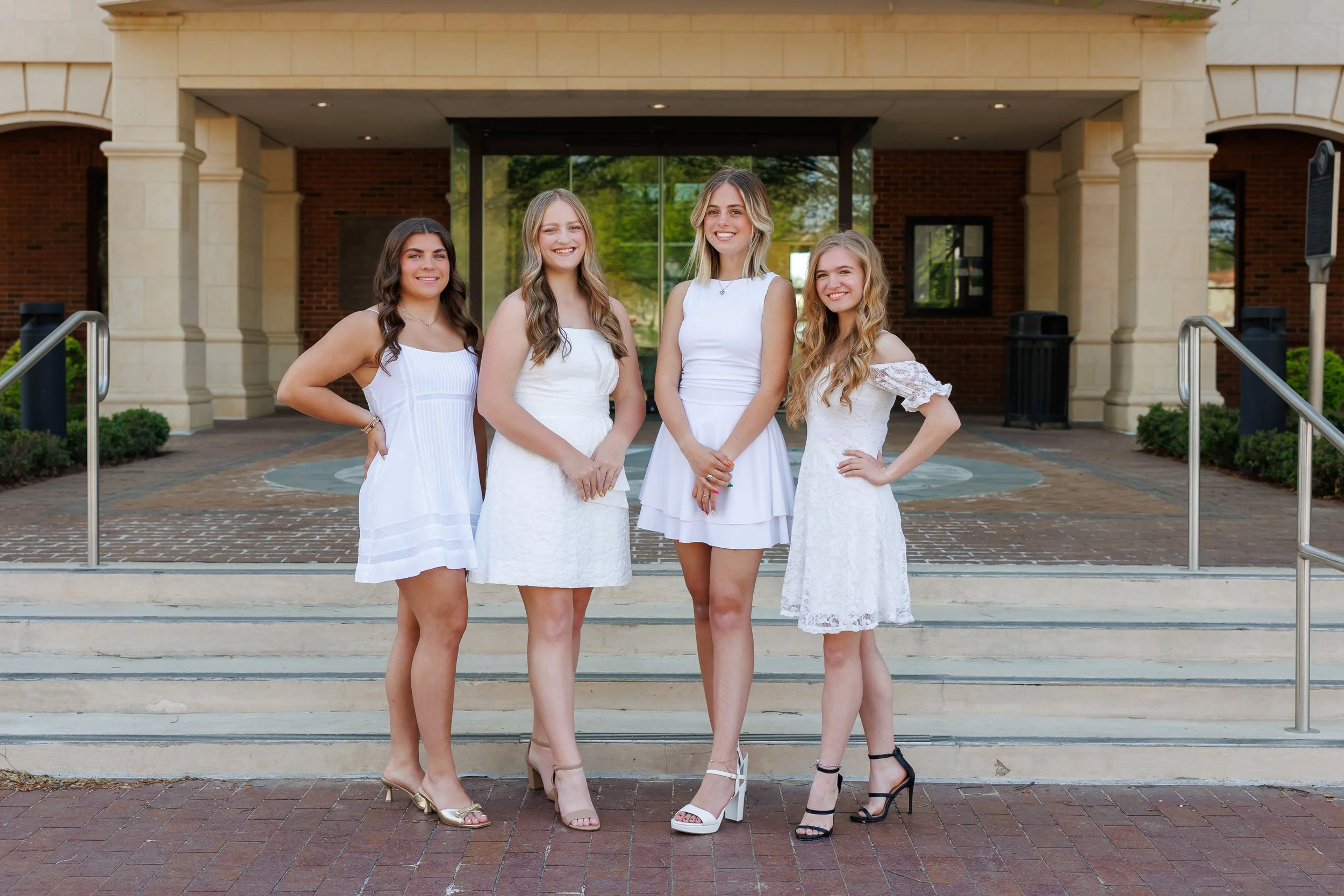 Four highschool girls in white dresses posing for senior class pictures on the steps of the Colleyville Library in Colleyville Texas
