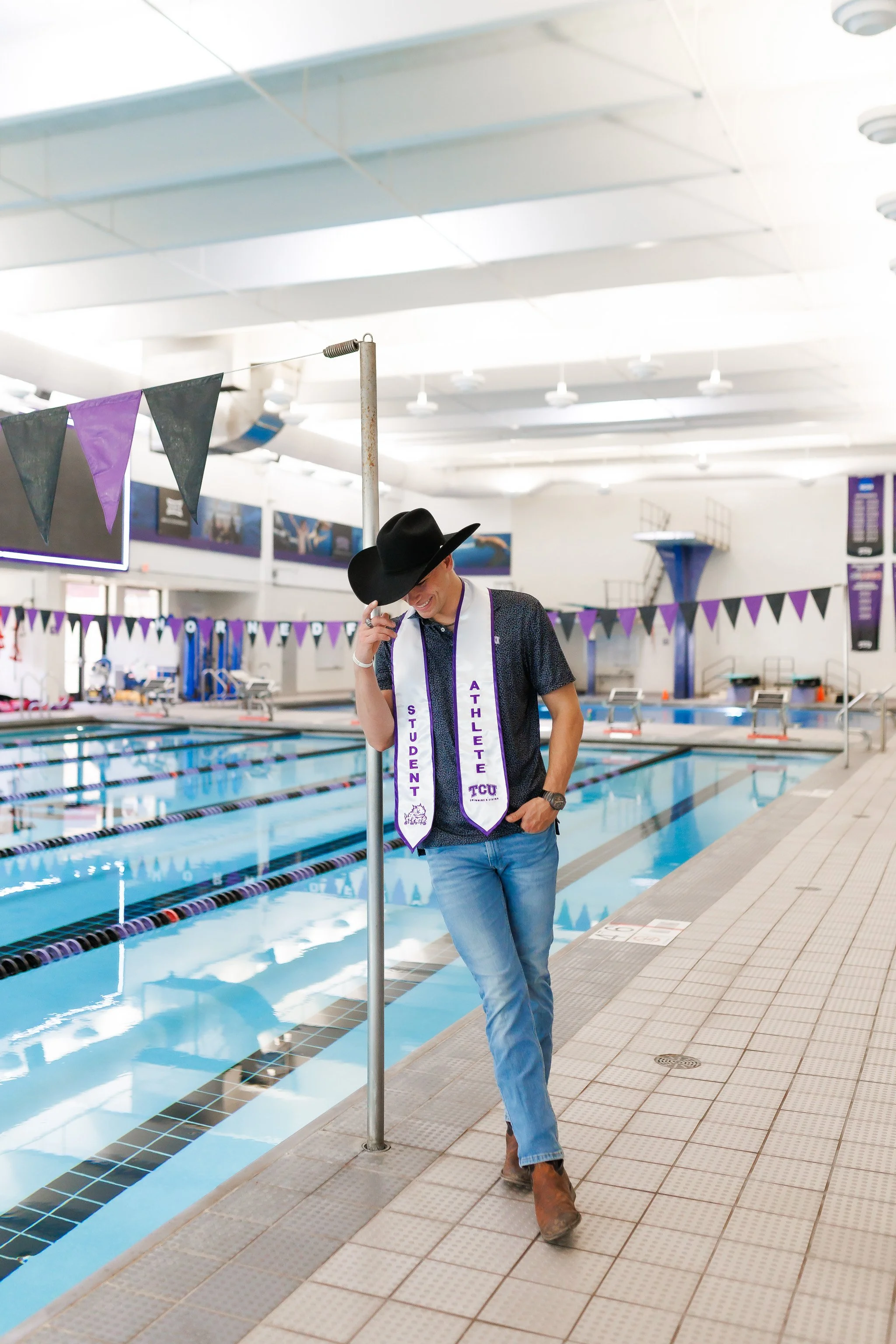 TCU Senior guy wearing a black cowboy hat and student athlete stole, while standing at the side of the pool at the University Recreation Center posing for senior pictures