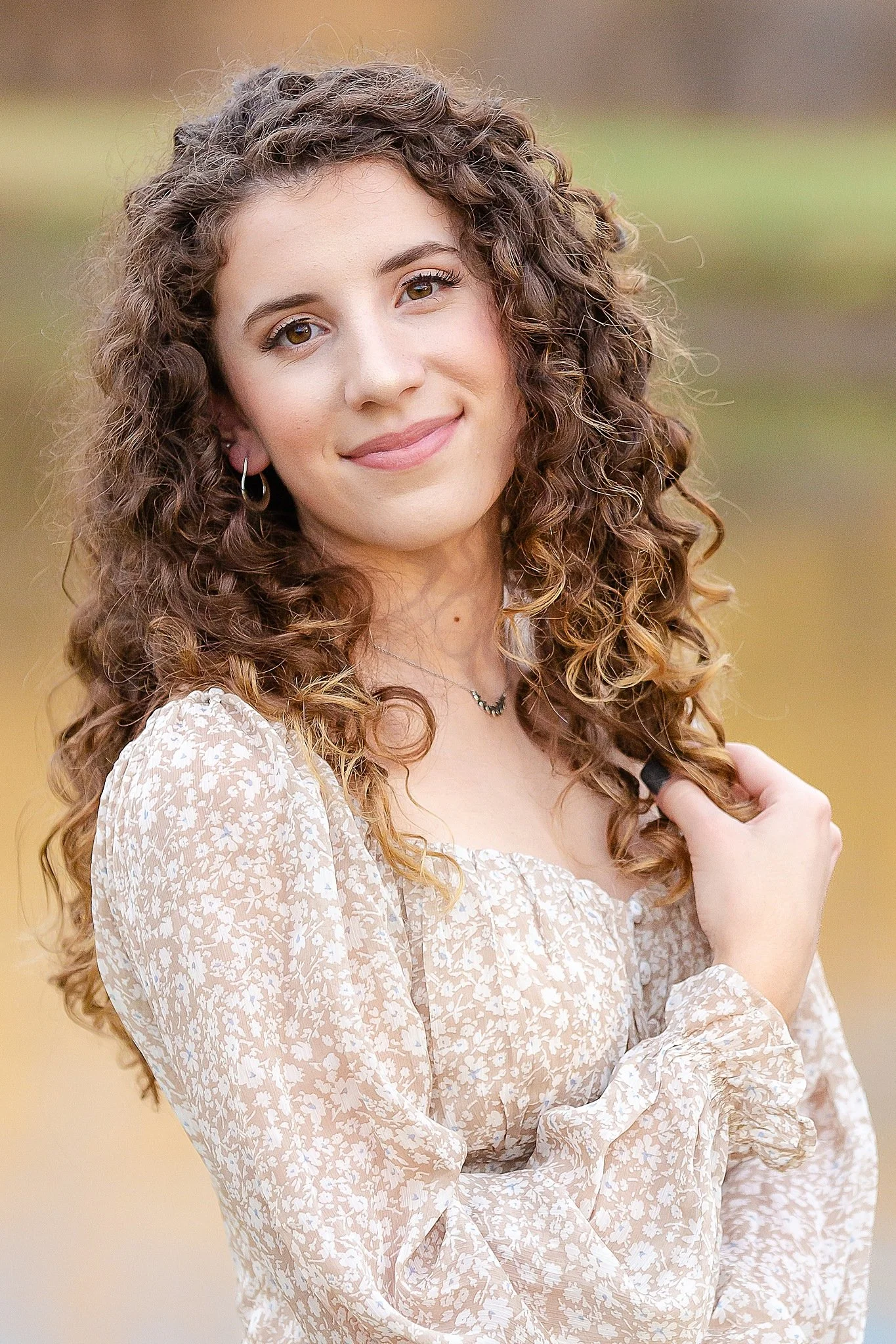 young lady with very curly long brown hair and big brown eyes wearing a light brown floral dress smiling at the camera for senior pictures at Colleyville Nature Center