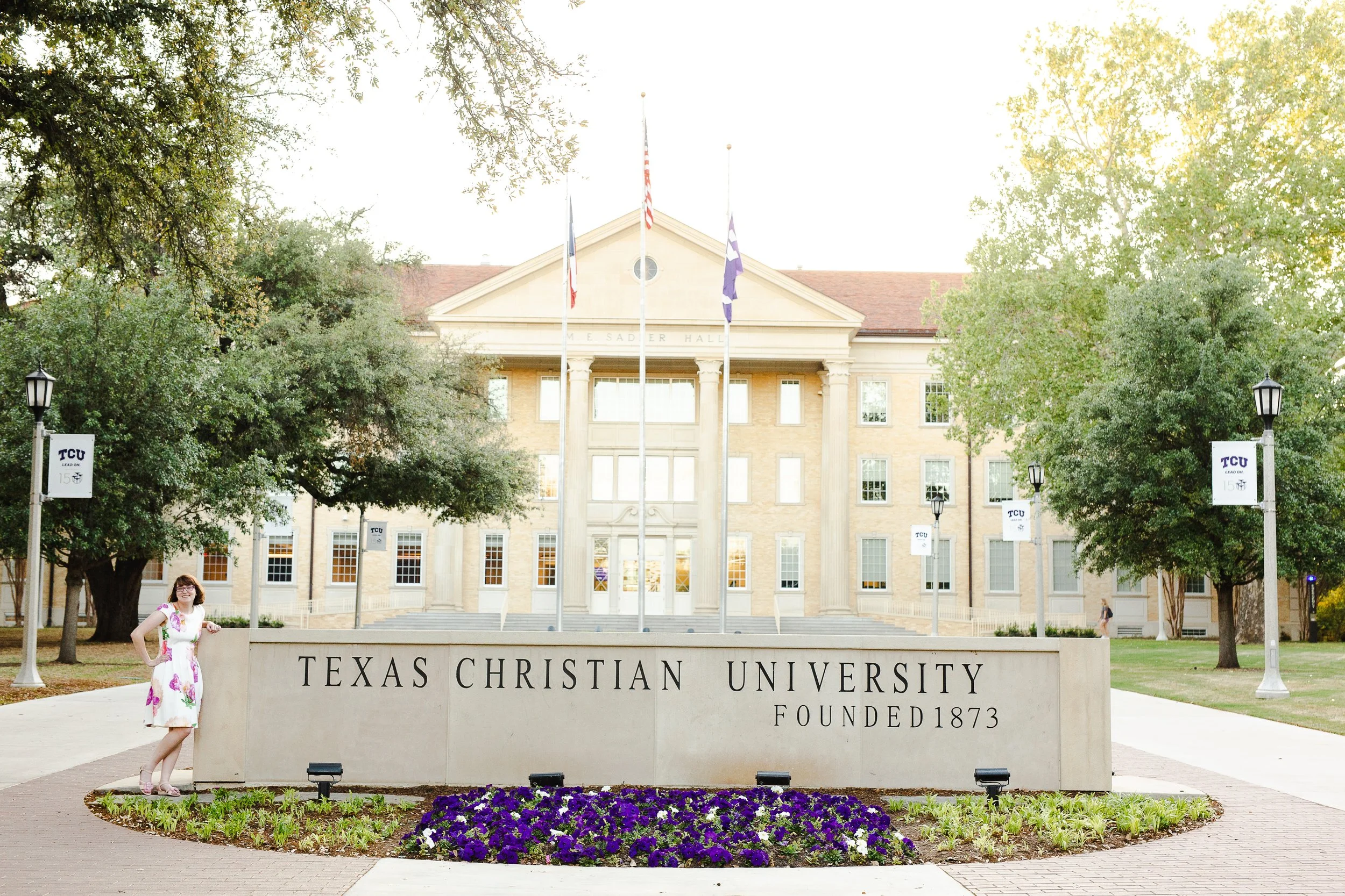 Graduate stands confidently beside the Texas Christian University sign in a floral dress, framed by lush greenery and historic Sadler Hall in the background