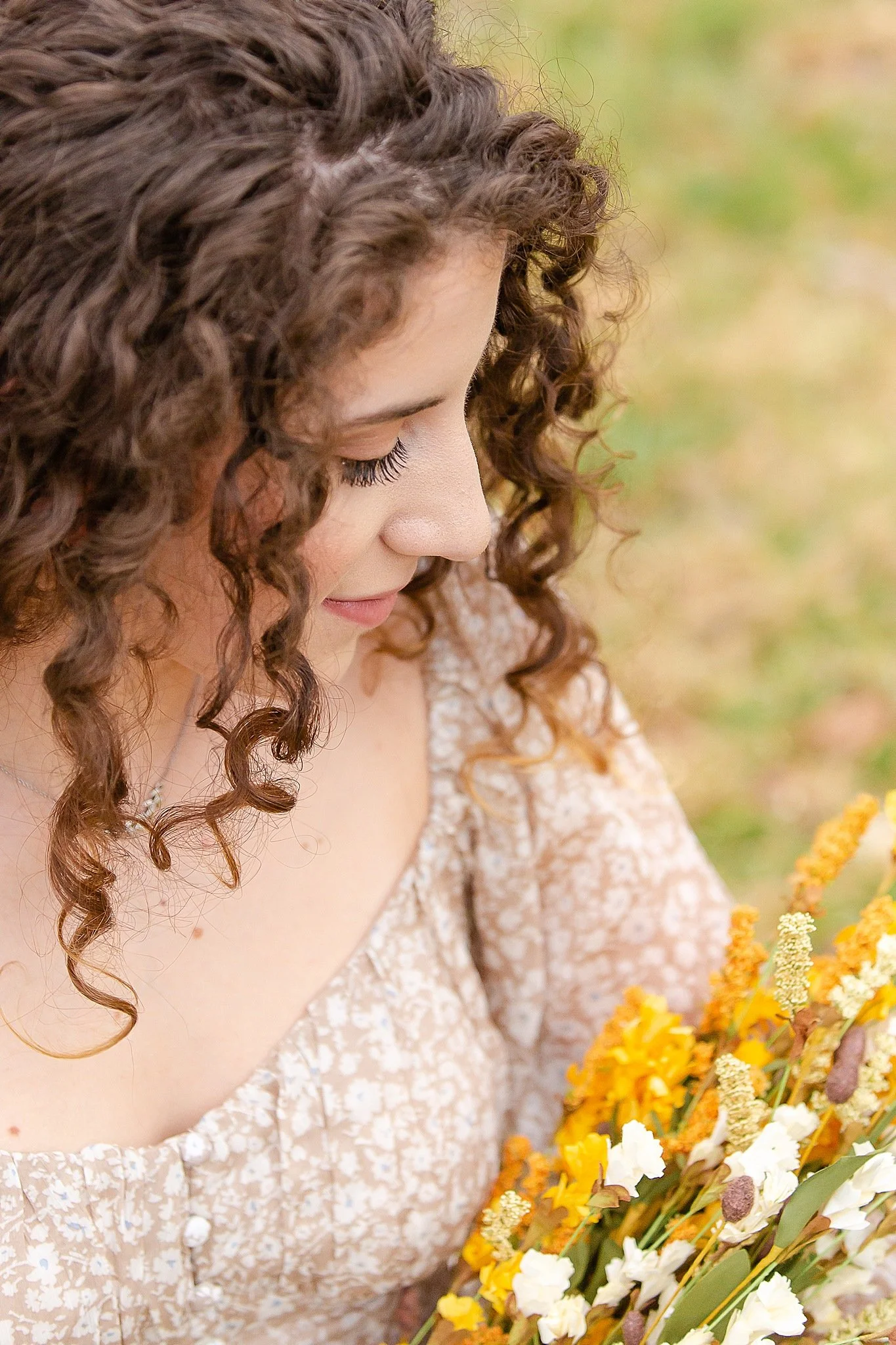 young lady with very curly long brown hair wearing a light brown floral dress holding a bouquet of dried flowers looking down at the flowers posing for senior pictures at Colleyville Nature Center