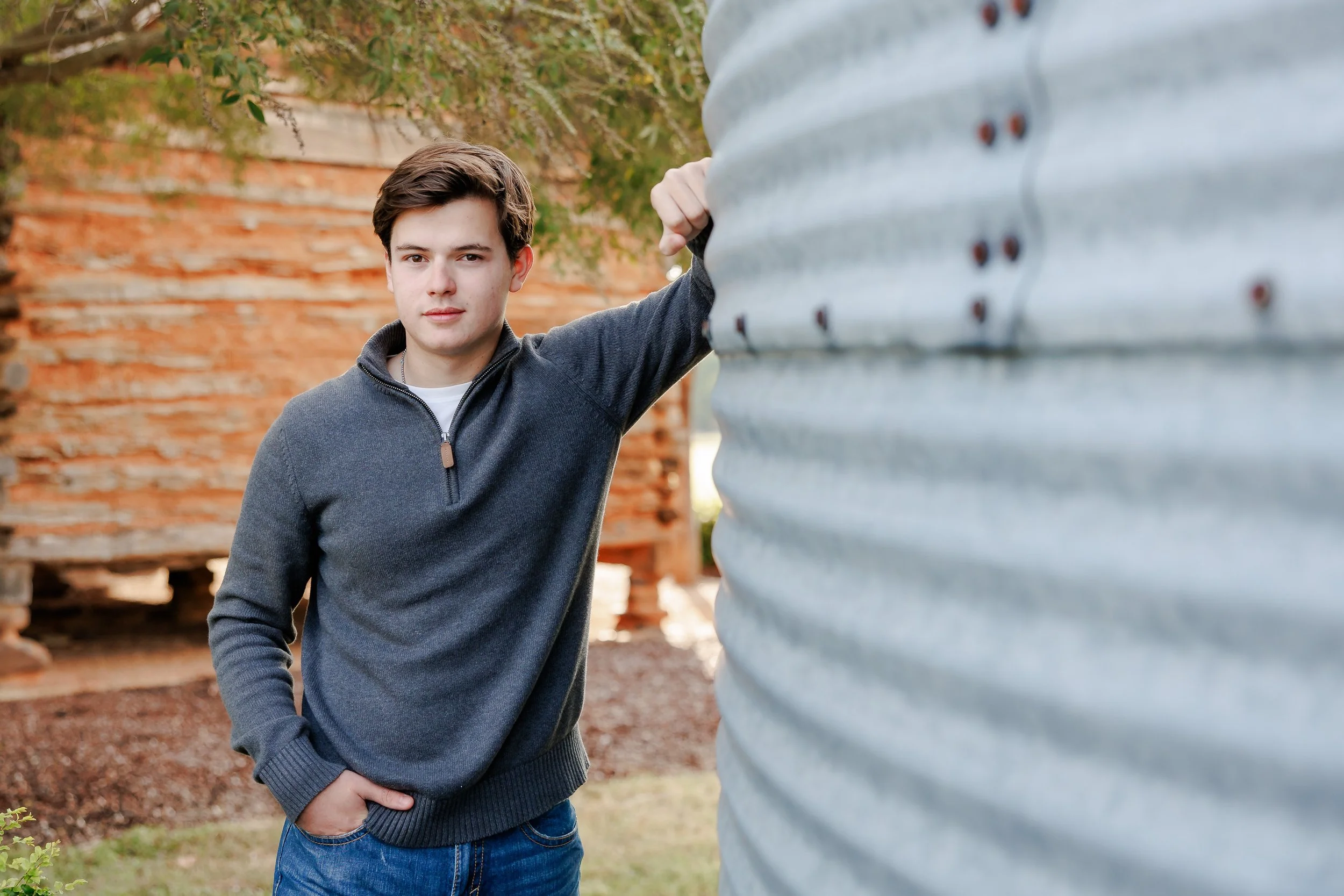 young man with wavy brown hair wearing a grey quarter zip and jeans smiling toward the camera with left arm resting above head onto a metal silo posing for professional senior portraits Colleyville Texas