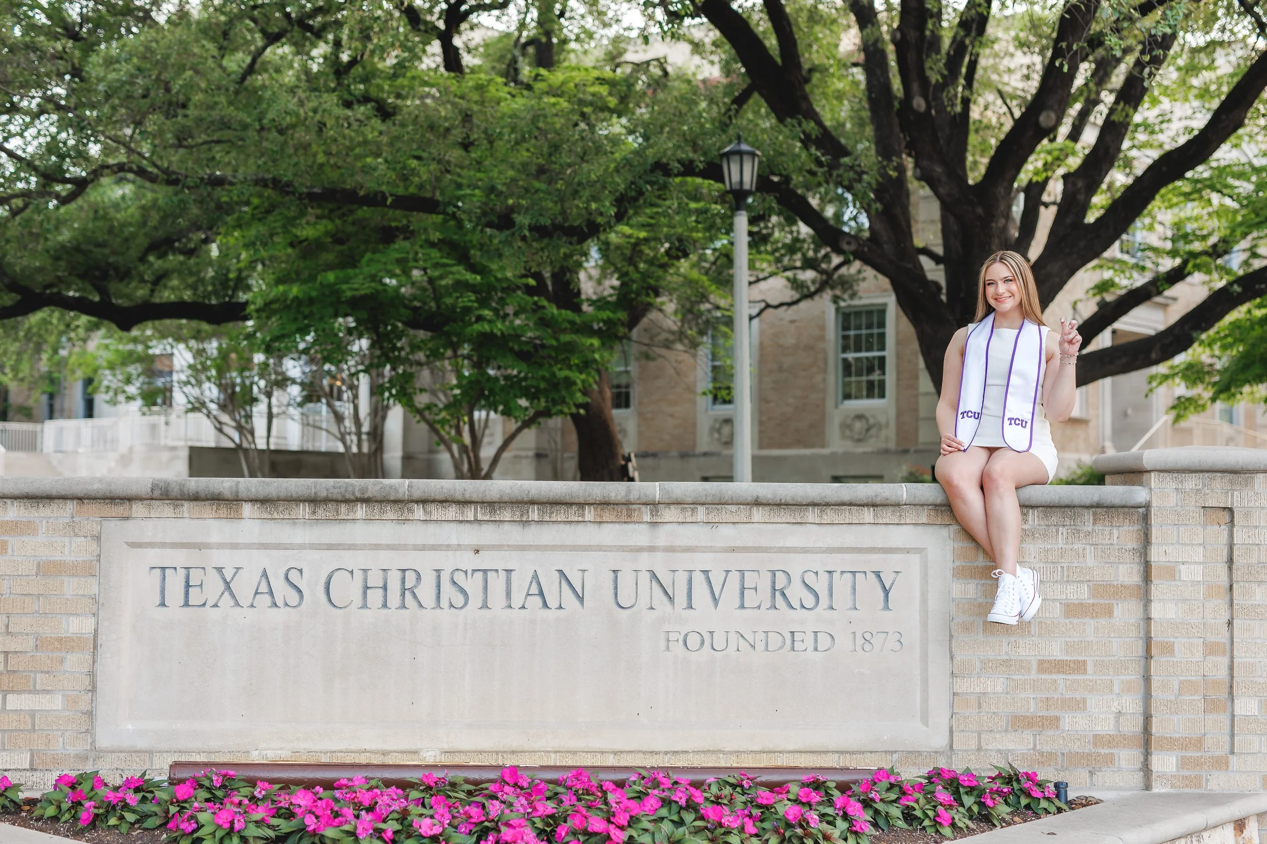 Senior girl wearing white dress sitting atop the stone Texas Christian University sign in front of Ed Landreth Hall at TCU posing for senior pictures