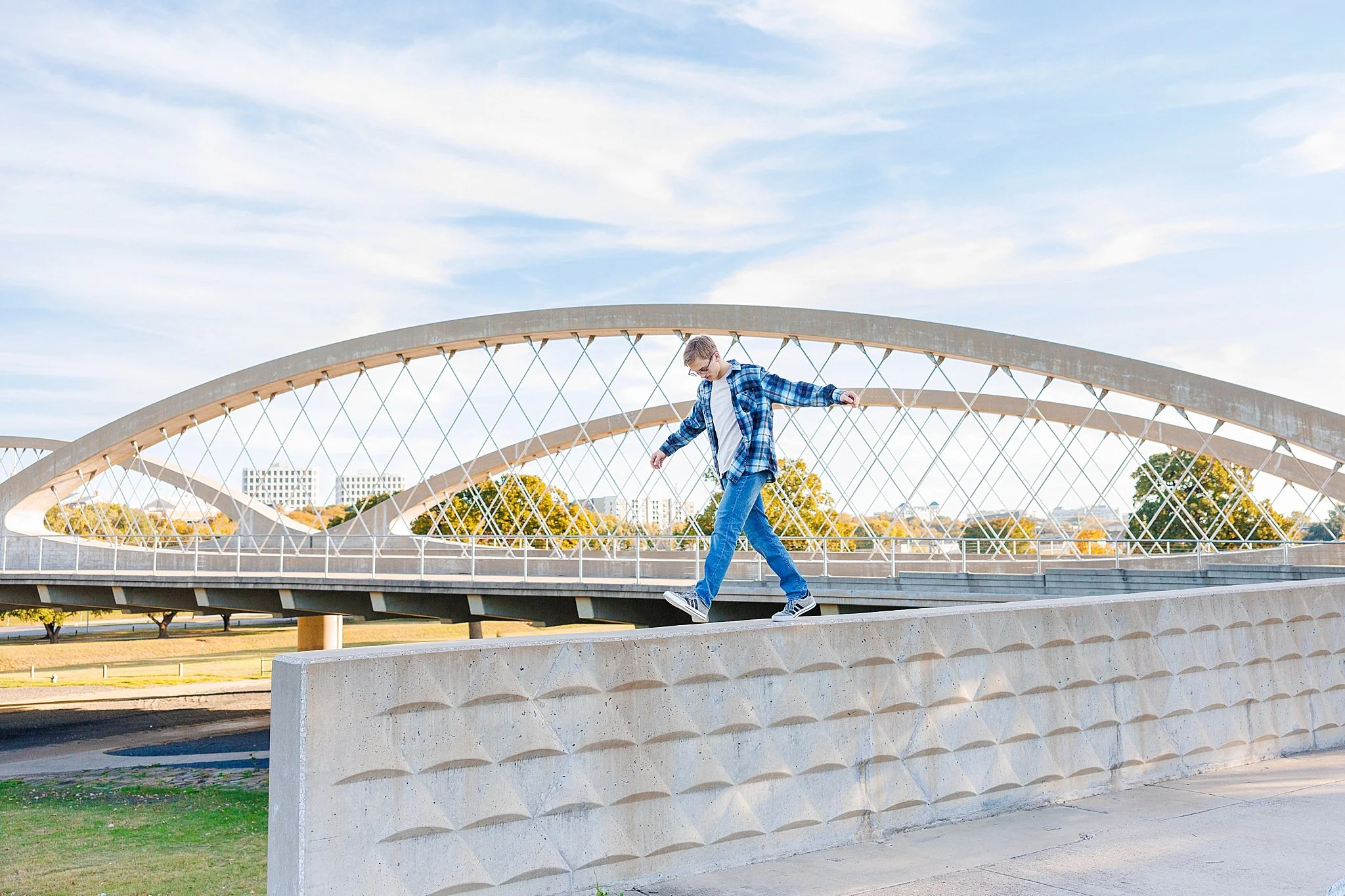 young man wearing jeans, tshirt, blue flannel shirt walking on top of concrete wall with arms spread wide in front of the 7th street bridge Fort Worth senior pictures