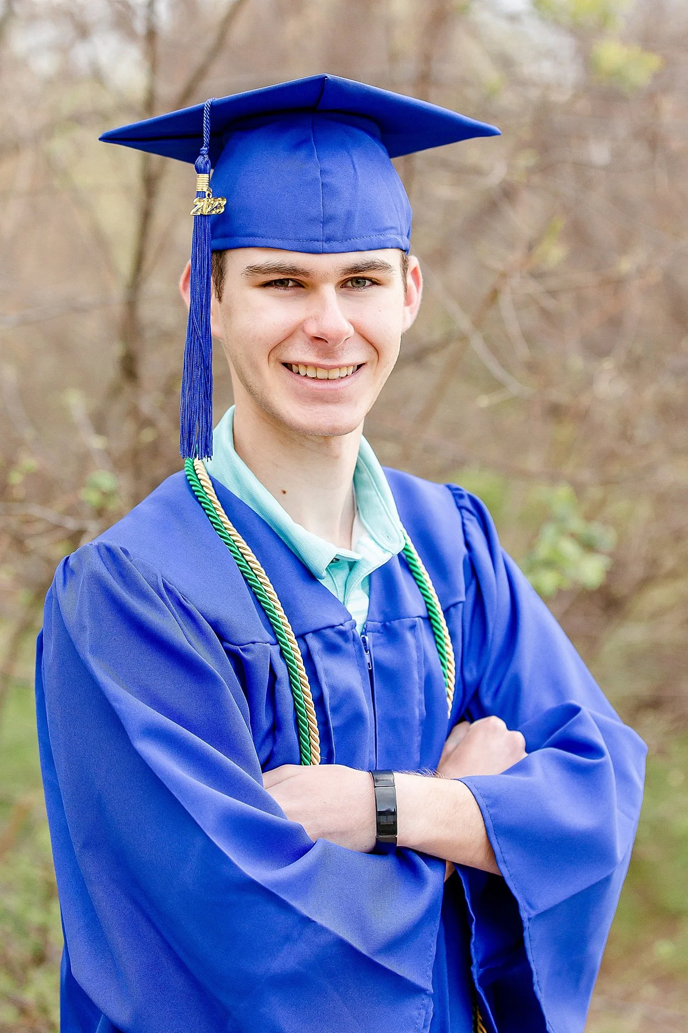 young man in blue graduation gown wearing a cap with tassel smiling at the camera posing for senior photos at Green Valley Park NRH TX