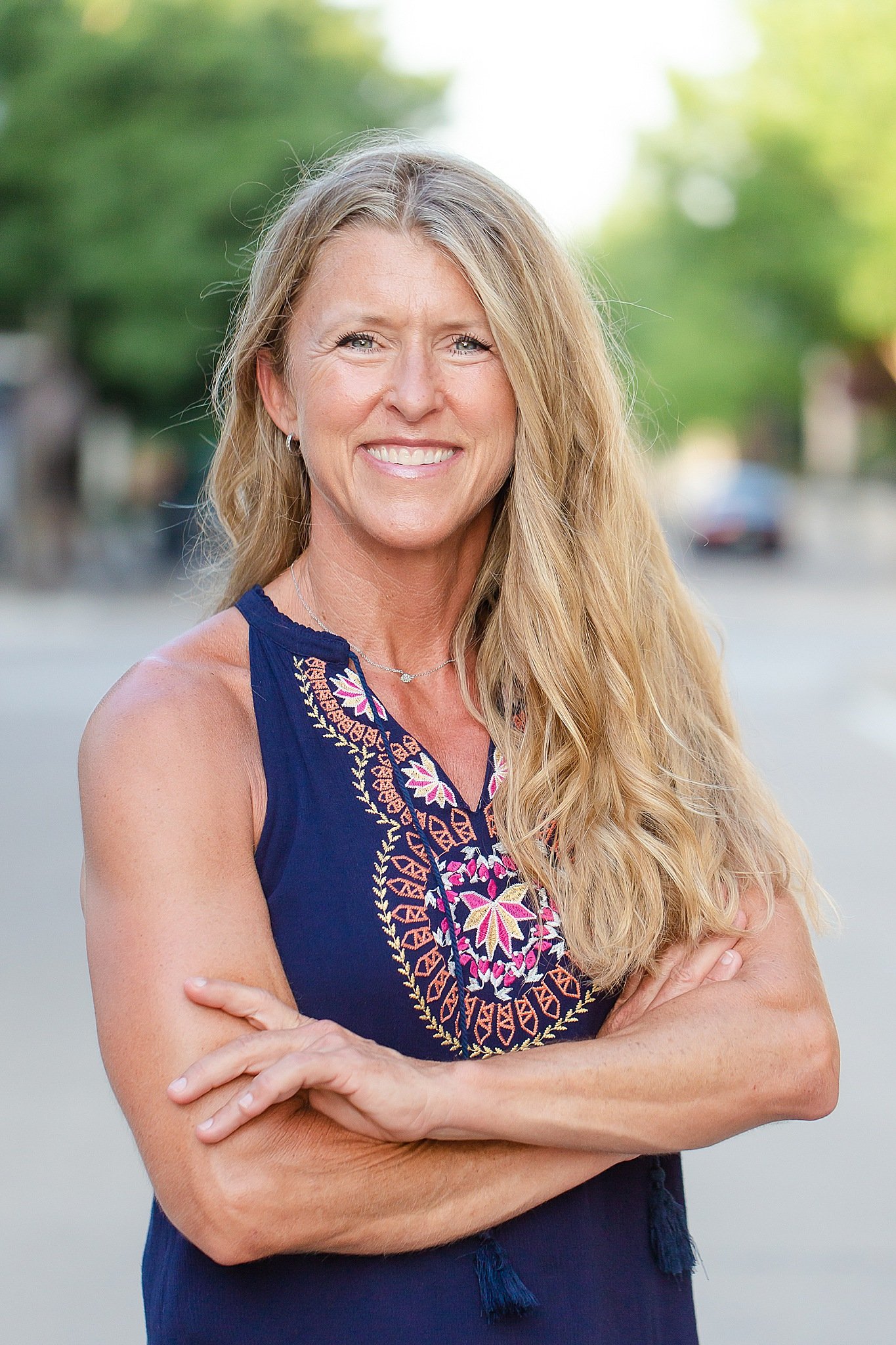 adult woman with long blond hair with beach waves wearing a dark blue sleeveless dress and statement earrings standing in a road with arms crossed posing for headshots at Colleyville Library