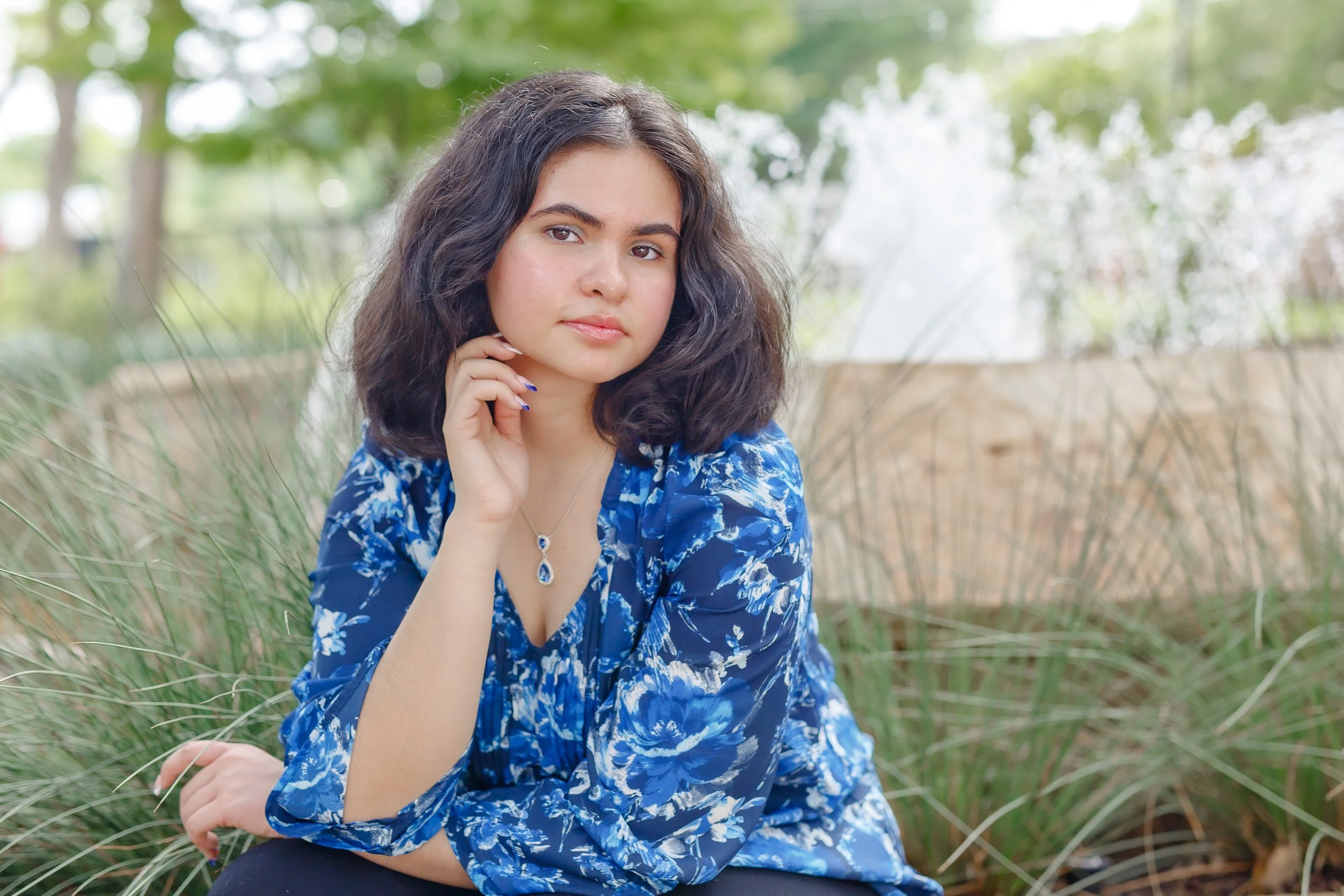 young lady with black wavy hair wearing a blue floral blouse resting her chin against her right hand staring somberly into the camera posing for professional portraits at Grapevine Botanical Gardens
