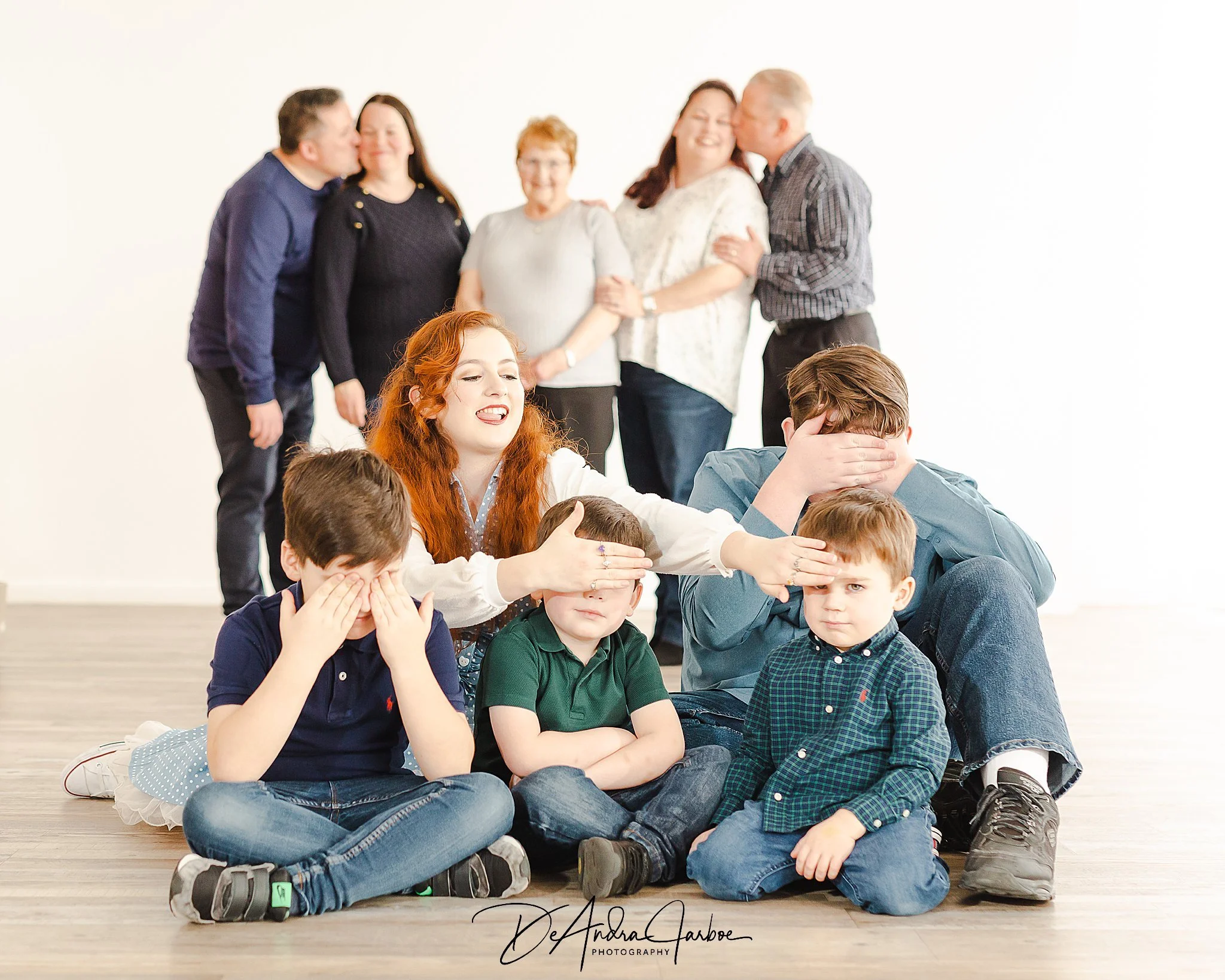 extended family portrait session of a family of ten with parents and grandparents blurred in the background and 5 grandkids in focus in the foreground. Parents are kissing kids are hiding their eyes.