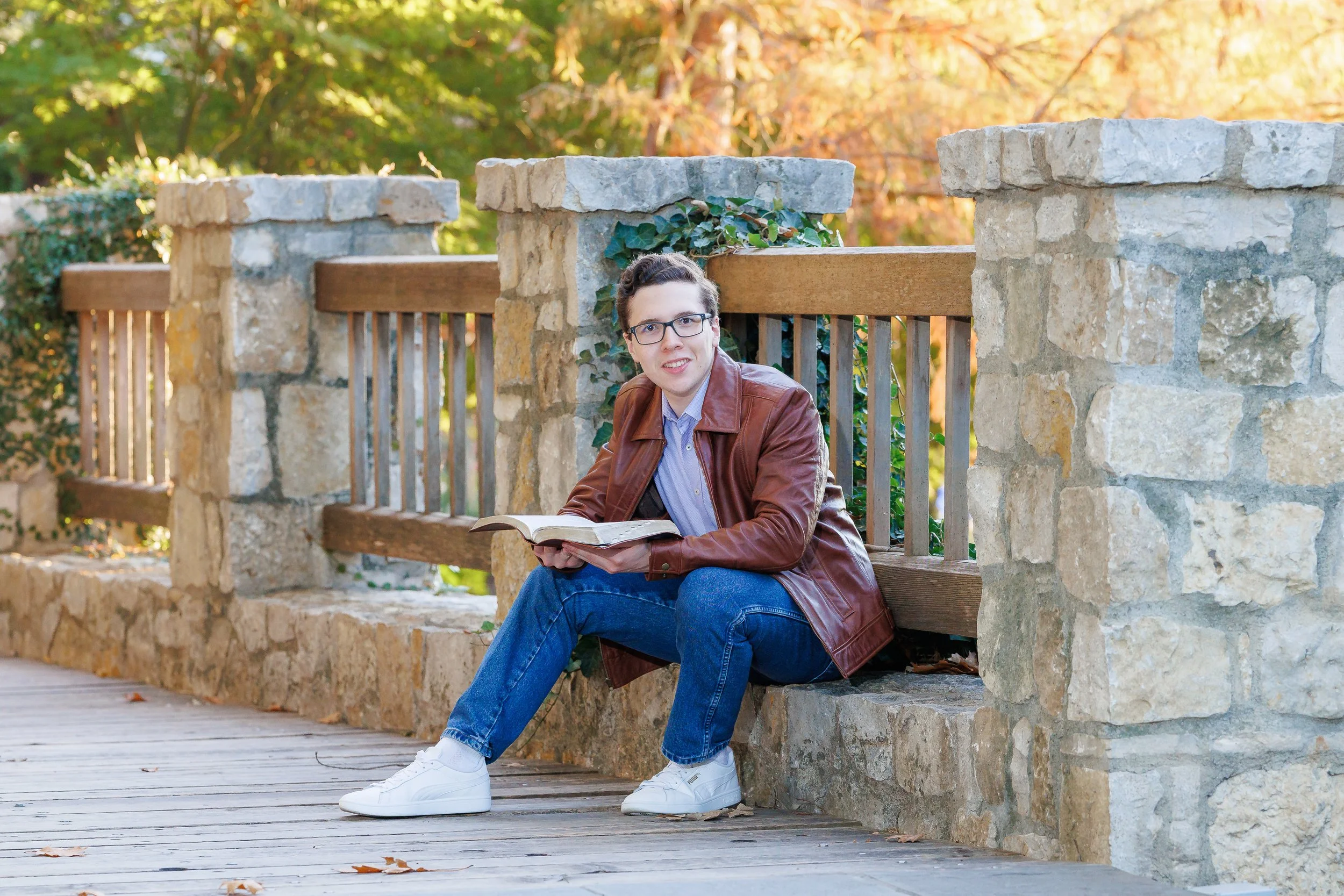 young man in a brown leather jacket, jeans, white tennis shoes holding an open Bible smiling at the camera posing for senior pictures at Dallas Arboretum
