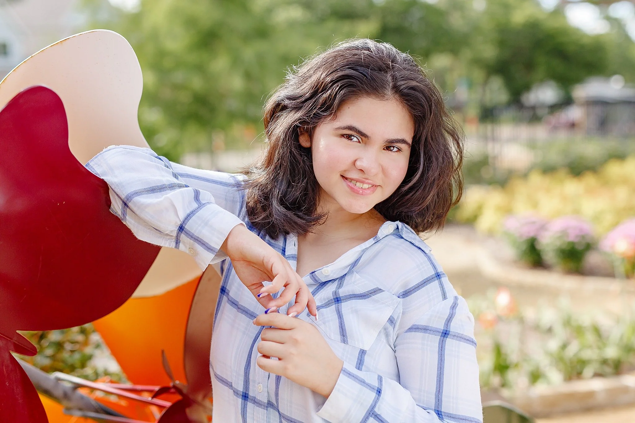 young lady with black wavy hair wearing a white blouse with blue stripes smiling at the camera  posing for senior pictures at grapevine botanical gardens