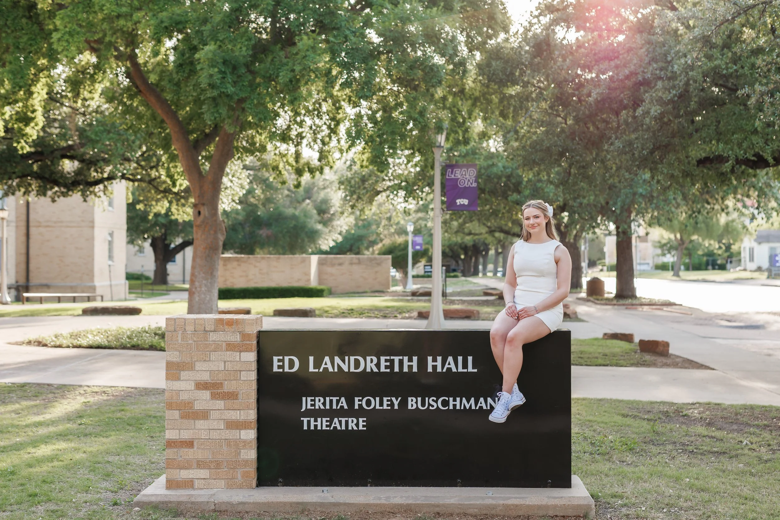Senior girl wearing white dress sitting on top of the sign at Ed Landreth Hall at TCU posing for senior pictures