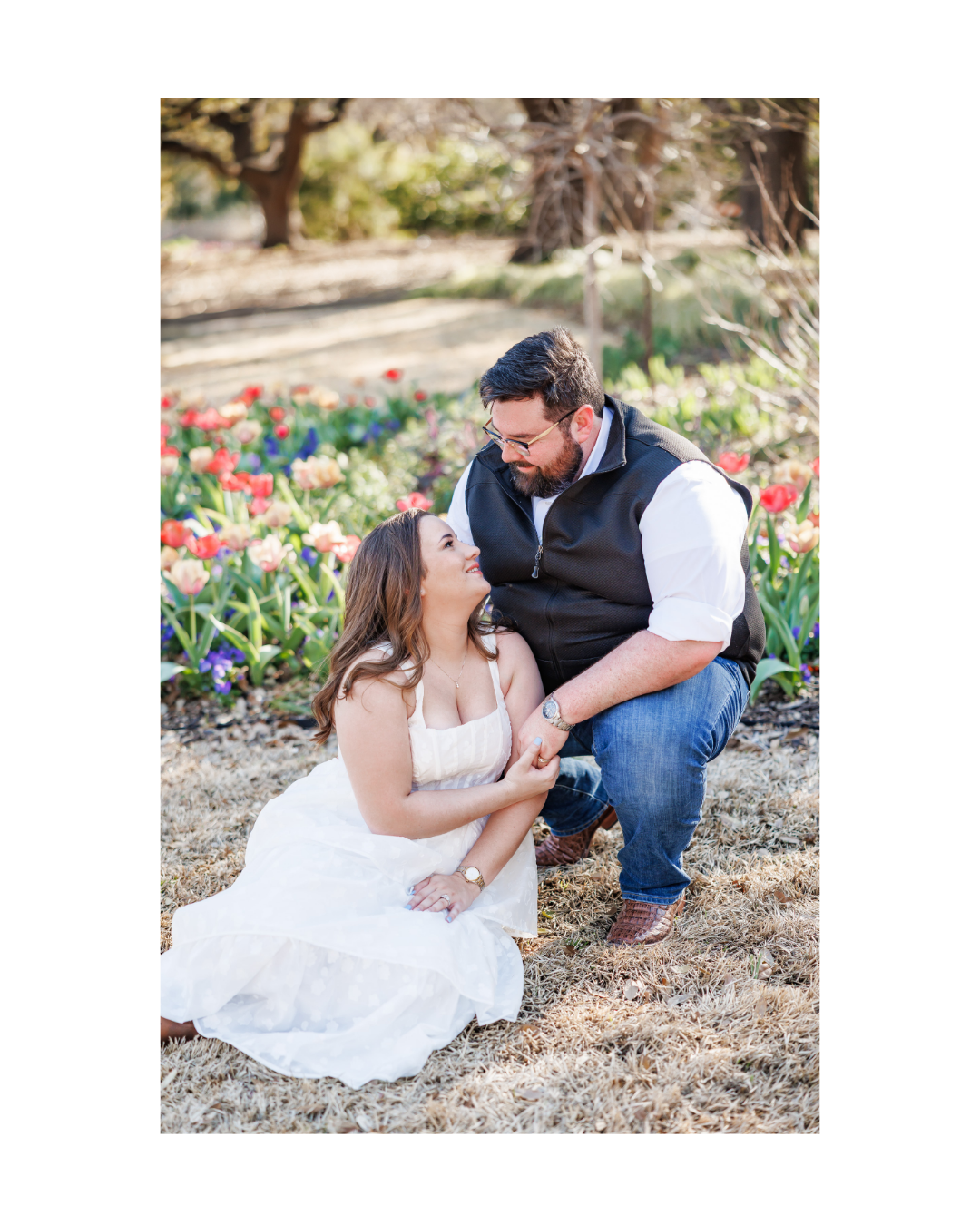 A couple in love outdoors among colorful flowers at Fort Worth Botanical Gardens, the woman sitting in a white dress and the man kneeling, holding her hand and smiling at her.