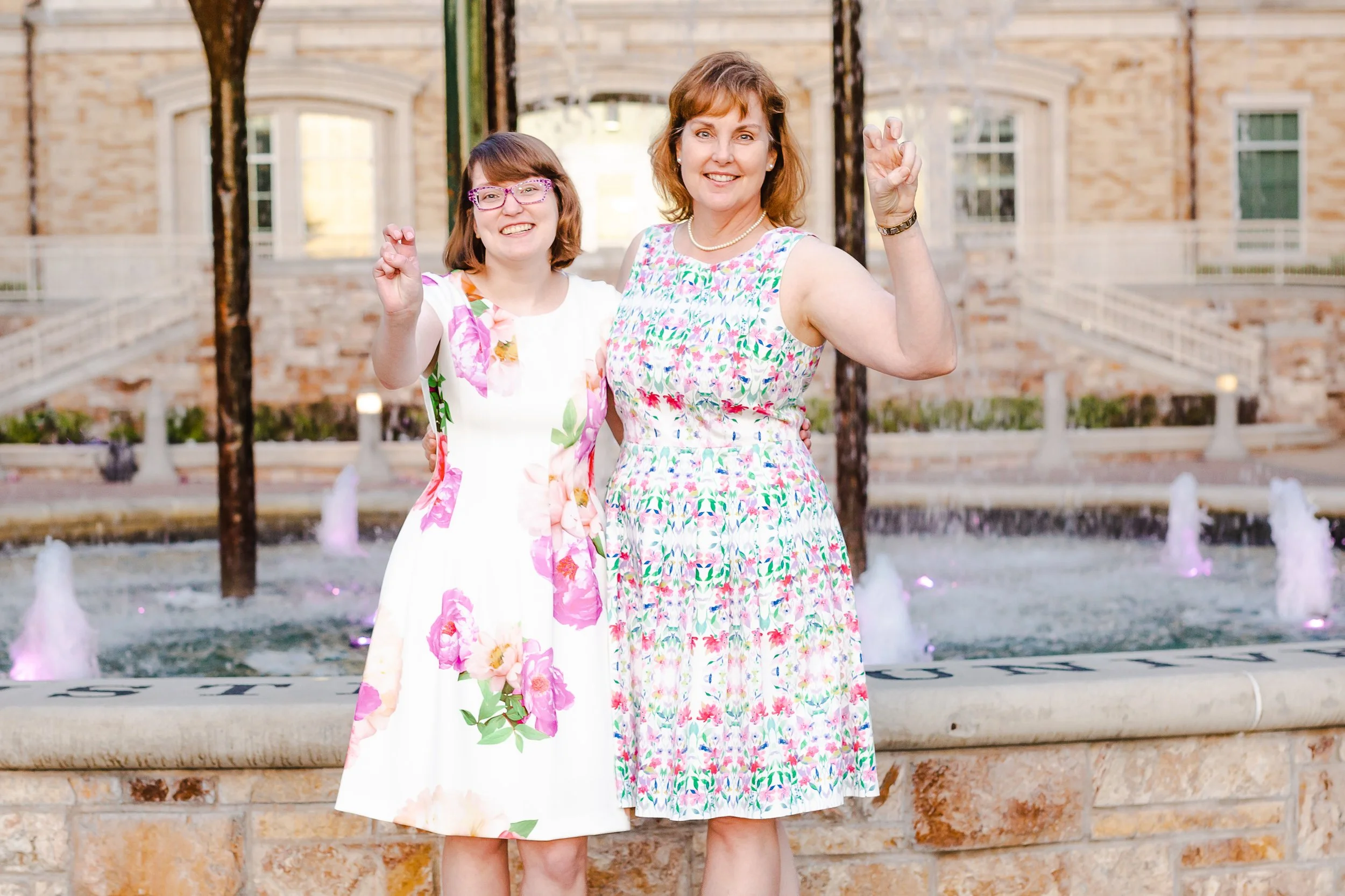 Graduate and her mother pose together in front of a fountain, both wearing floral dresses and holding up the Horned Frog hand sign, showcasing meaningful family-focused graduation photo pose ideas.