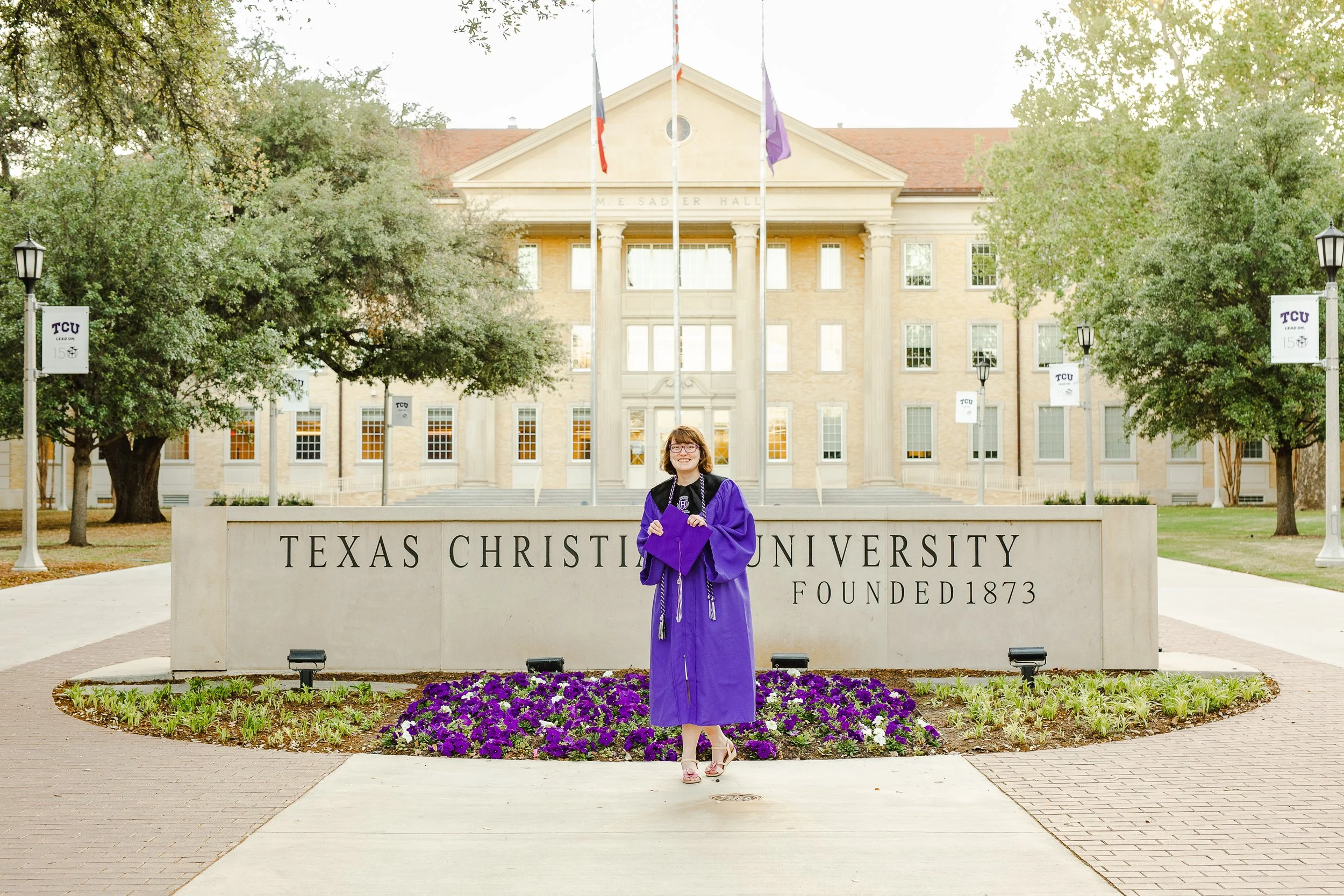 Graduate wearing a purple cap and gown poses in front of the Texas Christian University sign, holding her cap and smiling, offering timeless graduation photo pose ideas on campus.