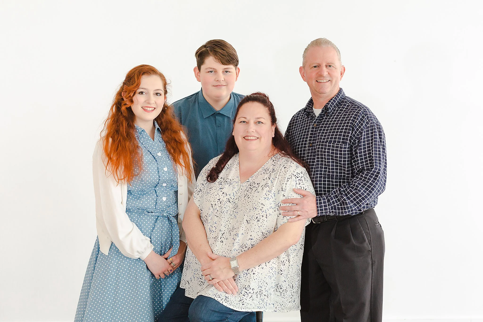 family of four posing for studio portraits. mom is siting on a stool with the kids and husband standing around her smiling at the camera