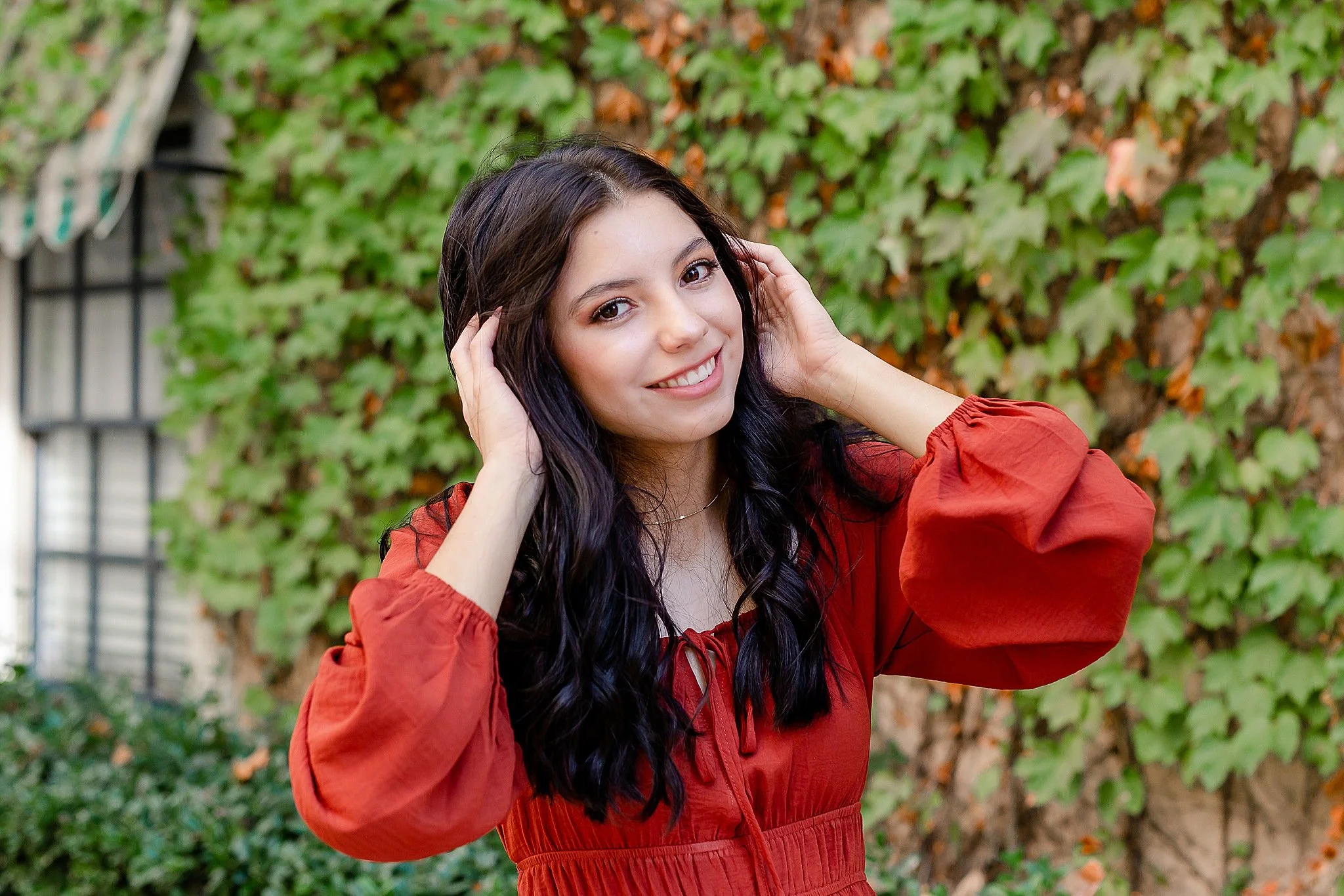 young lady with long black hair wearing a red long sleeve dress with both hands rustling her hair smiling at the camera  posing for senior pictures at Colleyville Library