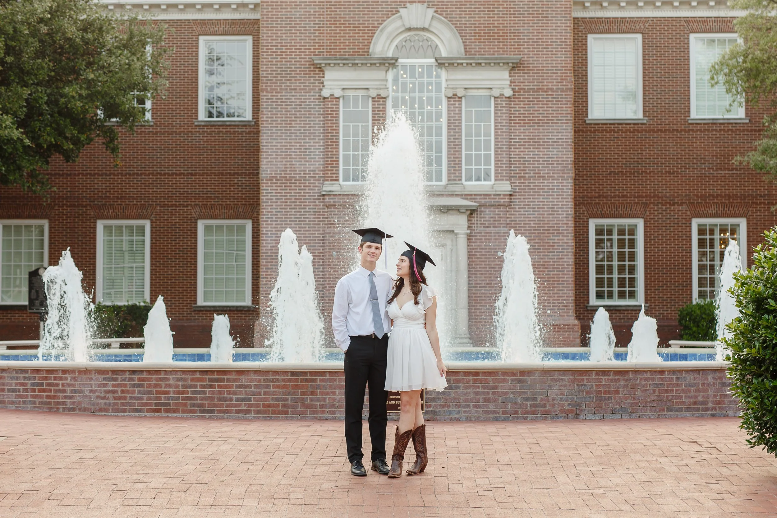 Couple posing in front of a fountain at Dallas Baptist University wearing graduation caps, with the woman in a white dress and cowboy boots and the man in a shirt and tie, captured for couples graduation photos by a Dallas photographer.