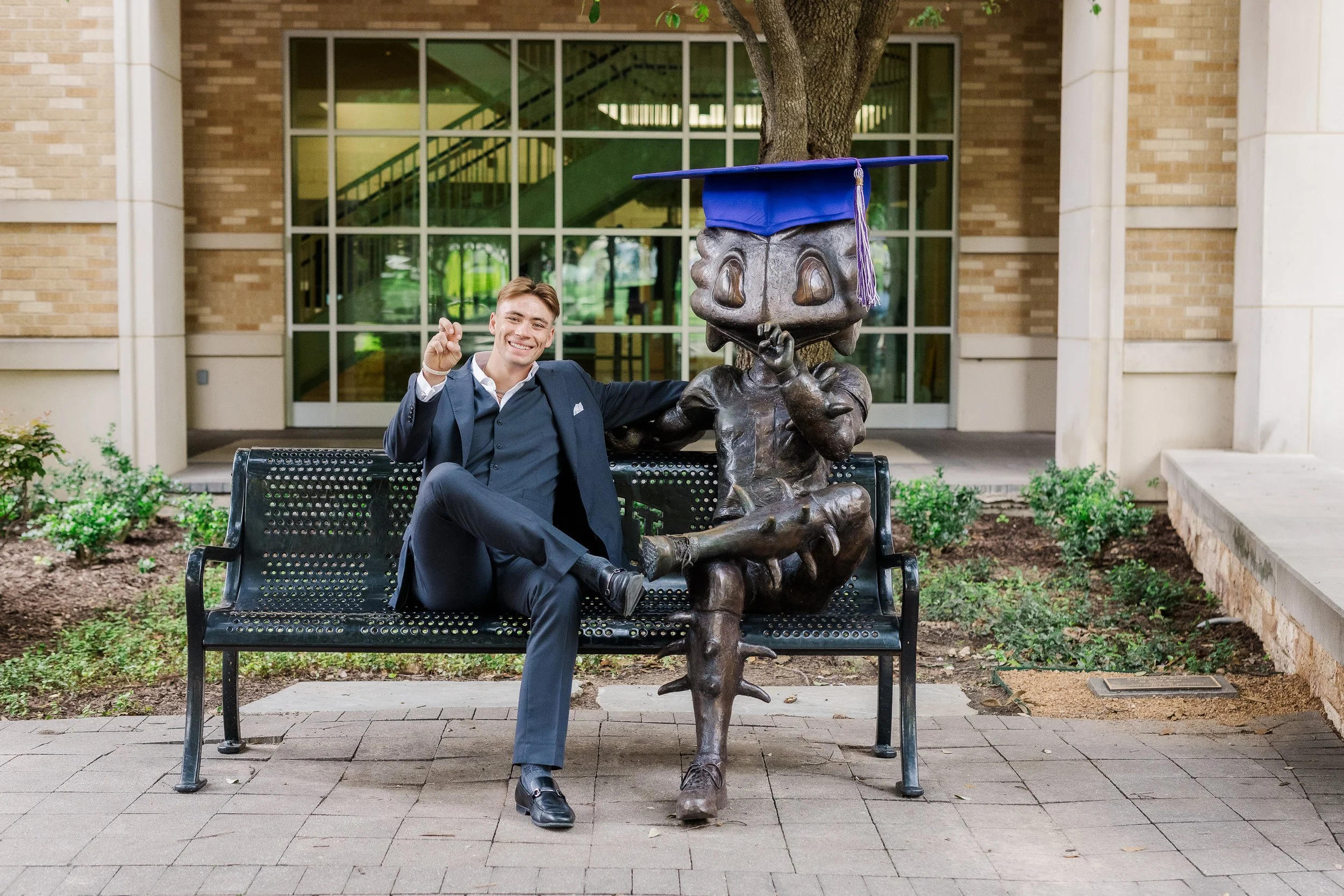 TCU senior guy wearing a grey suit sitting on a bench next to the TCU Superfrog statue in The Commons