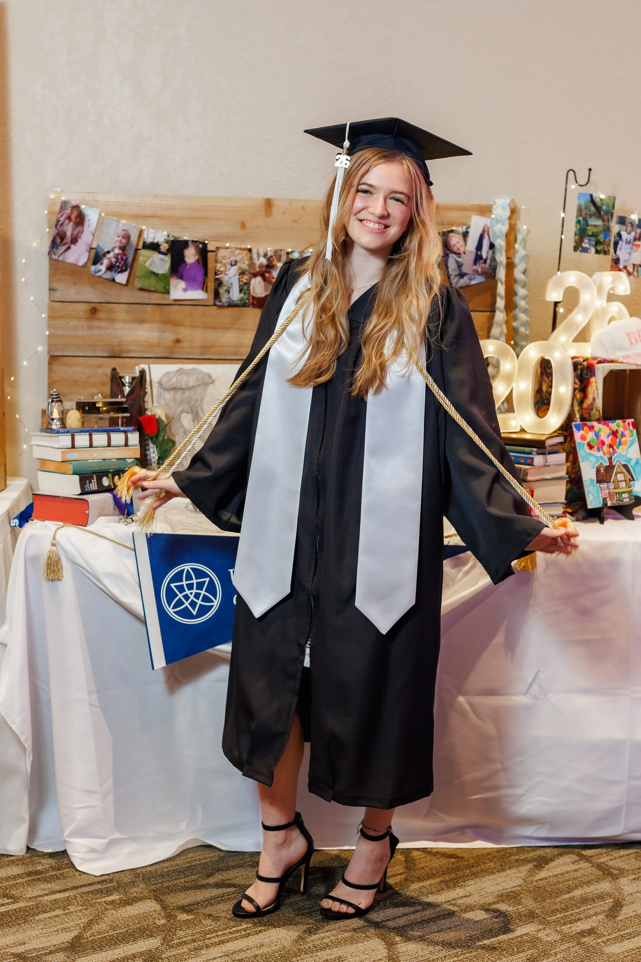highschool senior girl wearing black gown with white stole, gold honor cord and mortar board cap posing in front of a table with her awards and memories from school days. She's holding her honor cords out in celebration smiling at the camera.