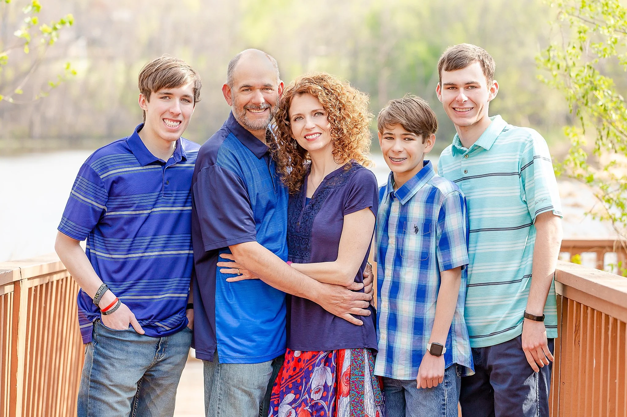 family of five smiling at the camera hugging joyfully while posing for senior pictures with one of their sons at Green Valley Park In NRH TX. Wearing shade of blue standing on dock with lake in background.