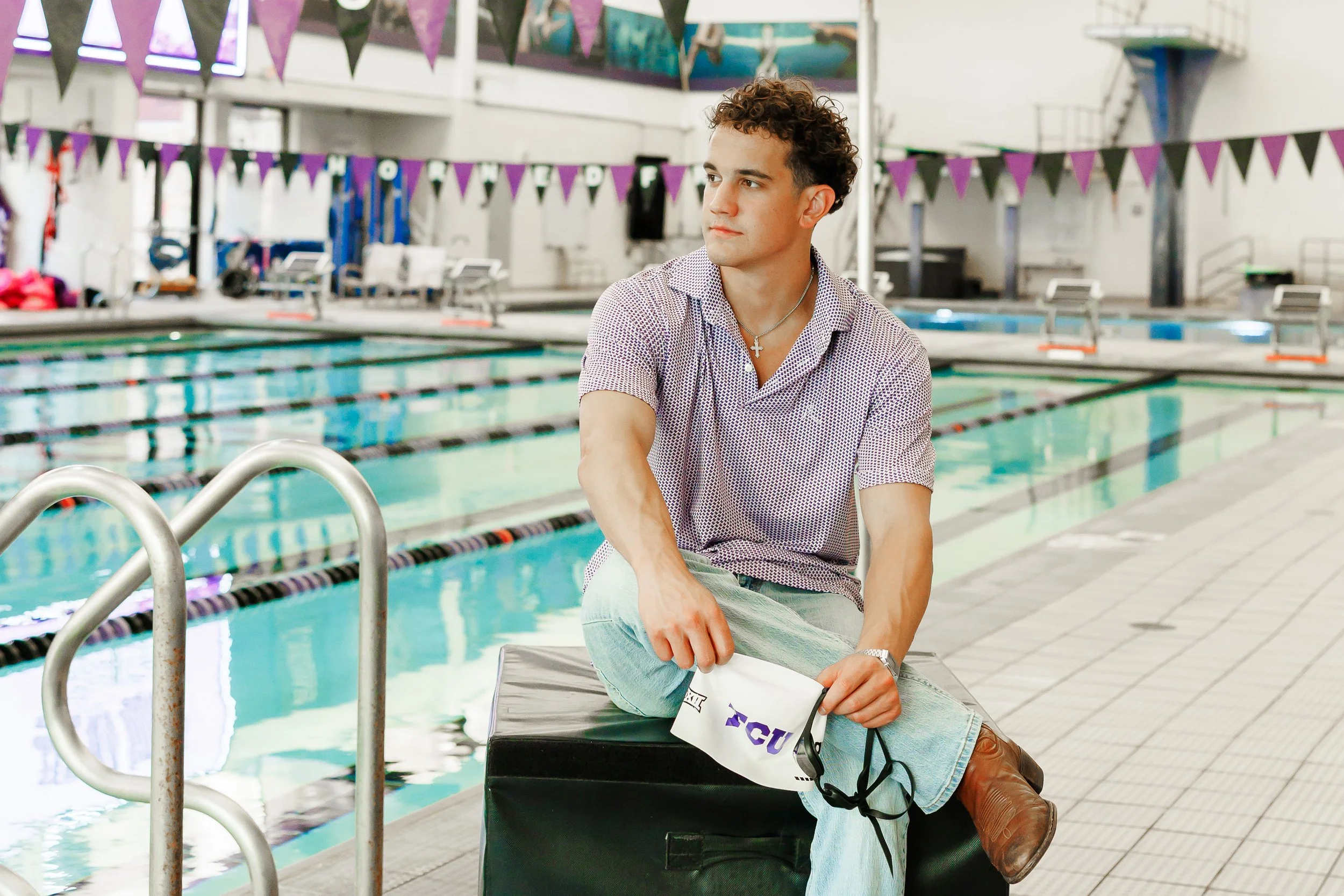 TCU Senior guy wearing purple shirt , holding a swim cap and goggles, sitting near the pool at the University Recreation Center posing for senior pictures