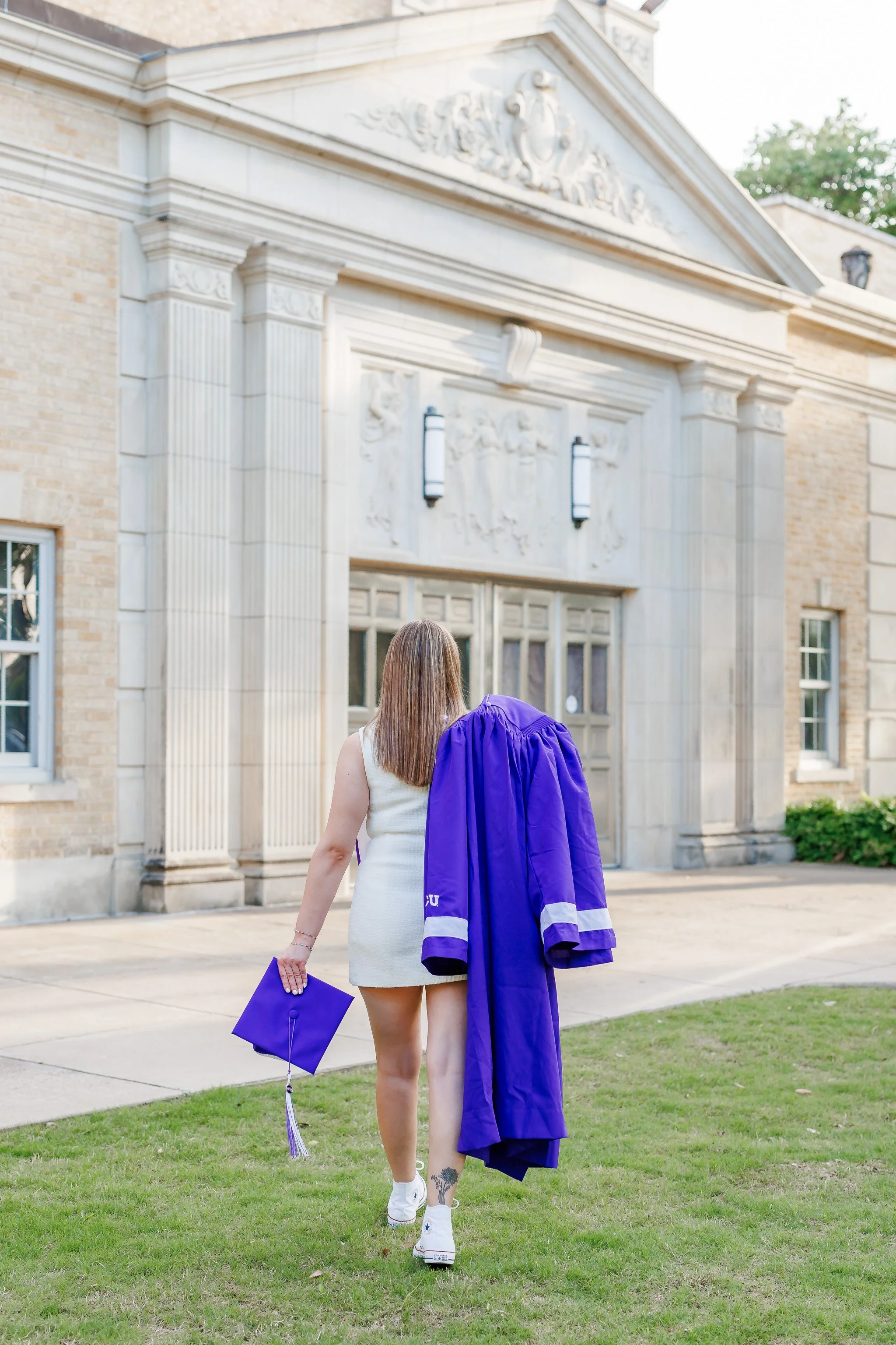 Senior girl wearing white dress walking with a graduation cap + gown in front of Ed Landreth Hall at TCU posing for senior pictures